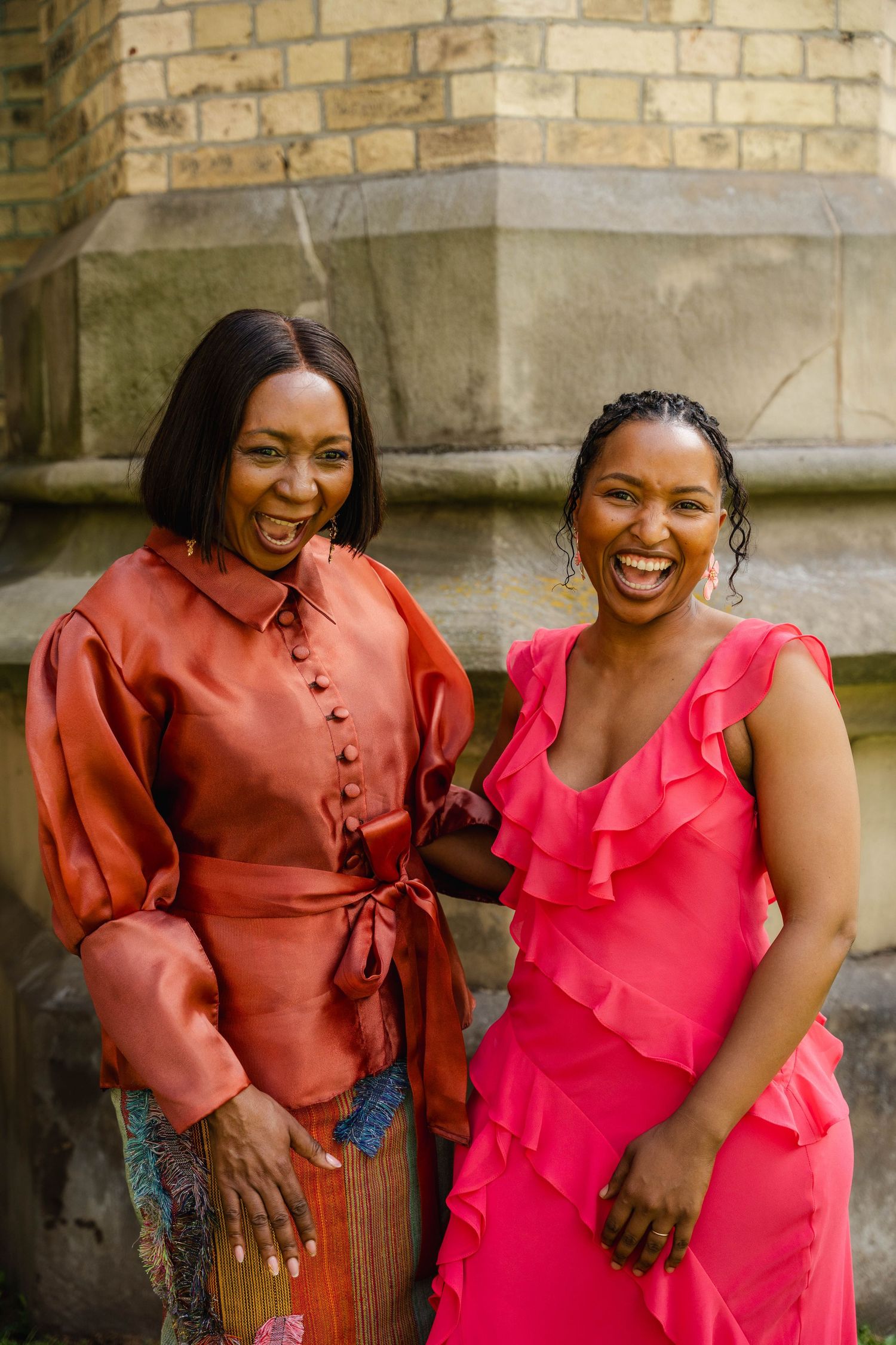 Two laughing women in a copper satin blouse and a bright pink ruffled dress pose together beside a stone church wall.