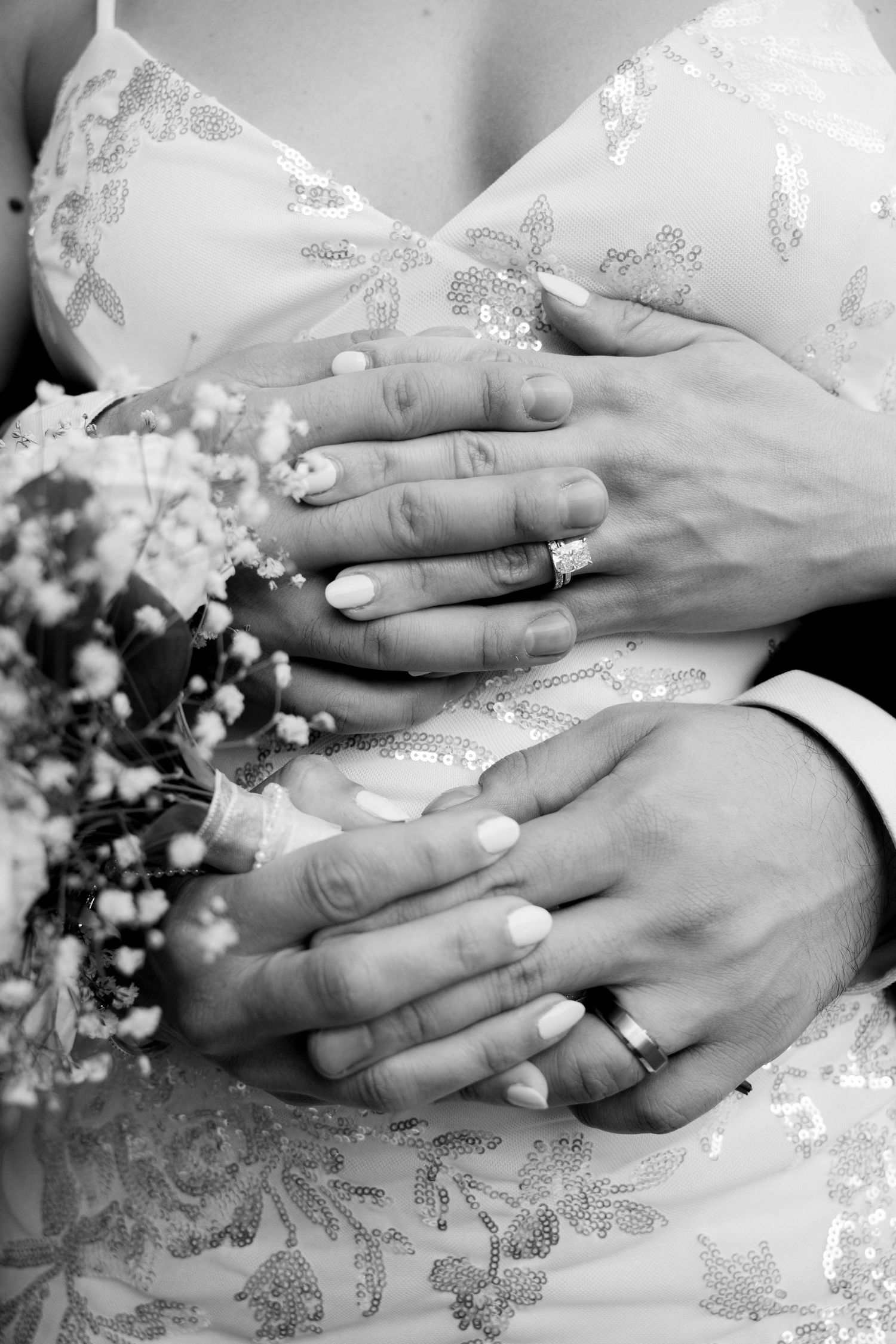 Black and white close-up of wedding rings and manicured hands resting on a sequined wedding dress next to a floral bouquet.