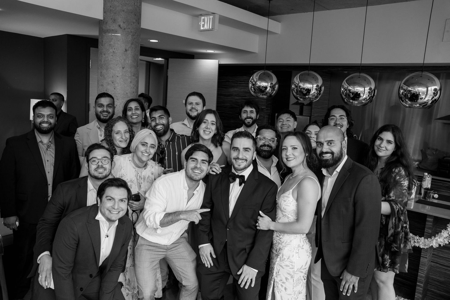 Black and white group portrait of a joyful bride and groom surrounded by smiling wedding guests in formal attire.