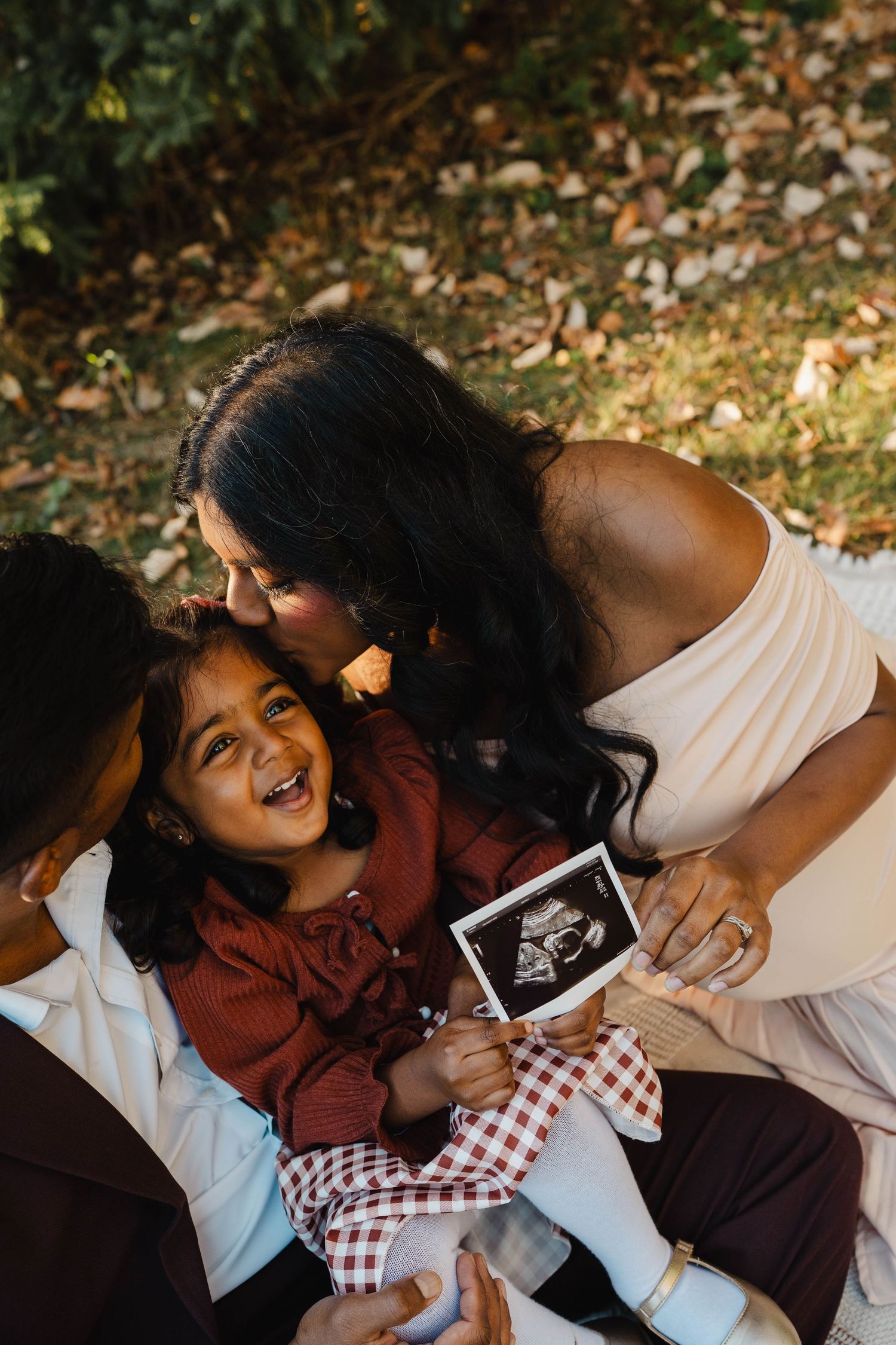 Smiling toddler girl holds an ultrasound photo while her pregnant mother kisses her cheek in a sunny outdoor family maternity portrait.