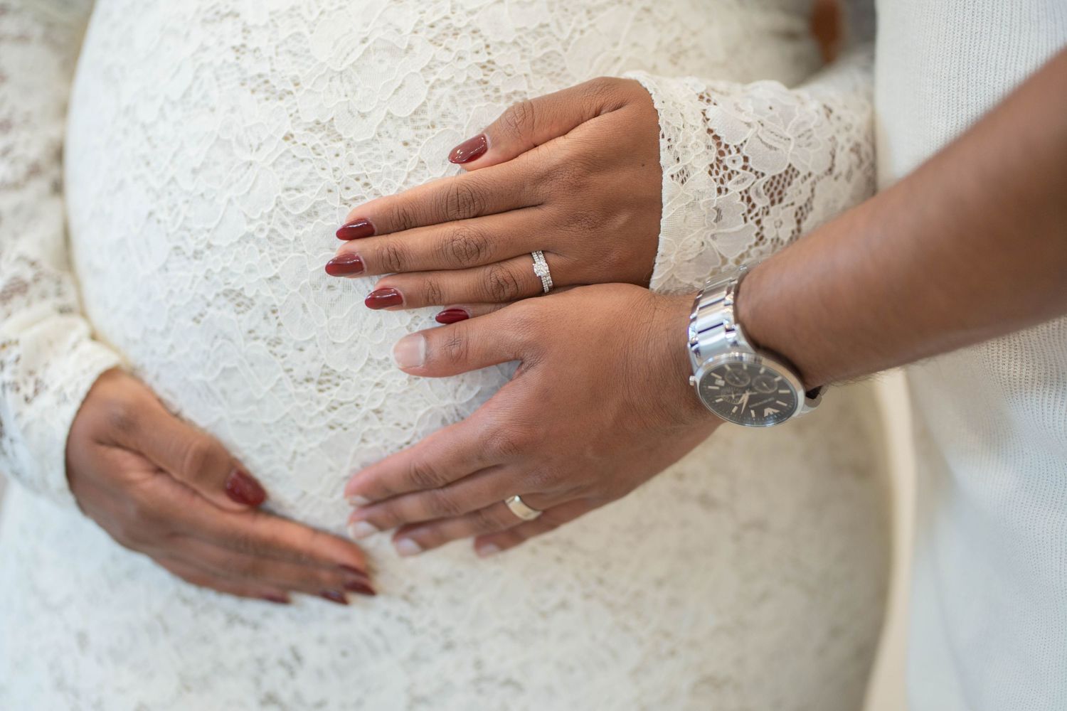 Close-up of a pregnant woman in a white lace dress and her partner holding her belly, showing rings and a silver watch.