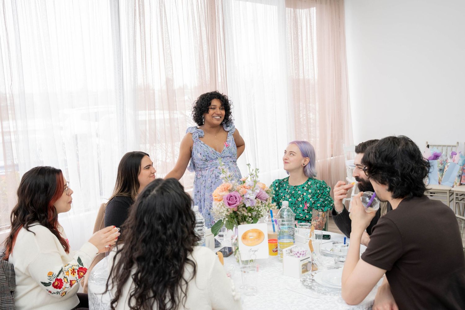 A woman in a blue dress stands smiling behind a table of seated guests with a floral centerpiece at a shower.