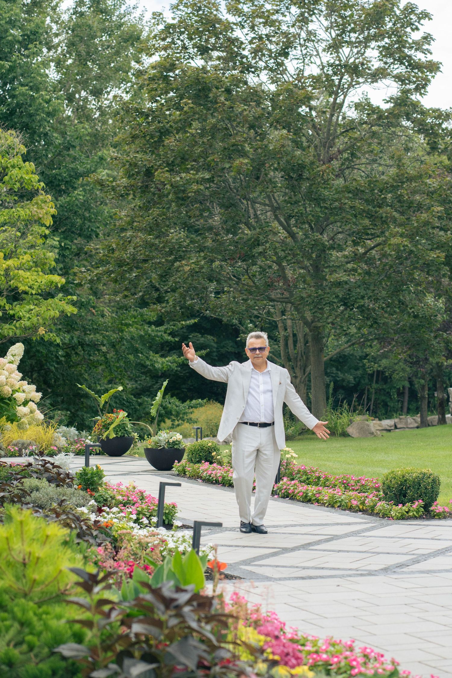 Smiling man in a light suit and sunglasses stands with arms outstretched on a stone path lined with colorful flowers.