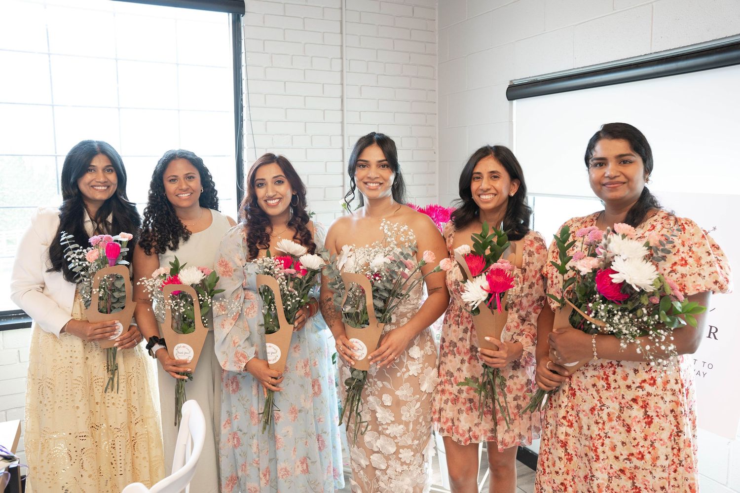 Six women in floral dresses pose together at a bridal shower, each holding a fresh flower bouquet wrapped in kraft paper.