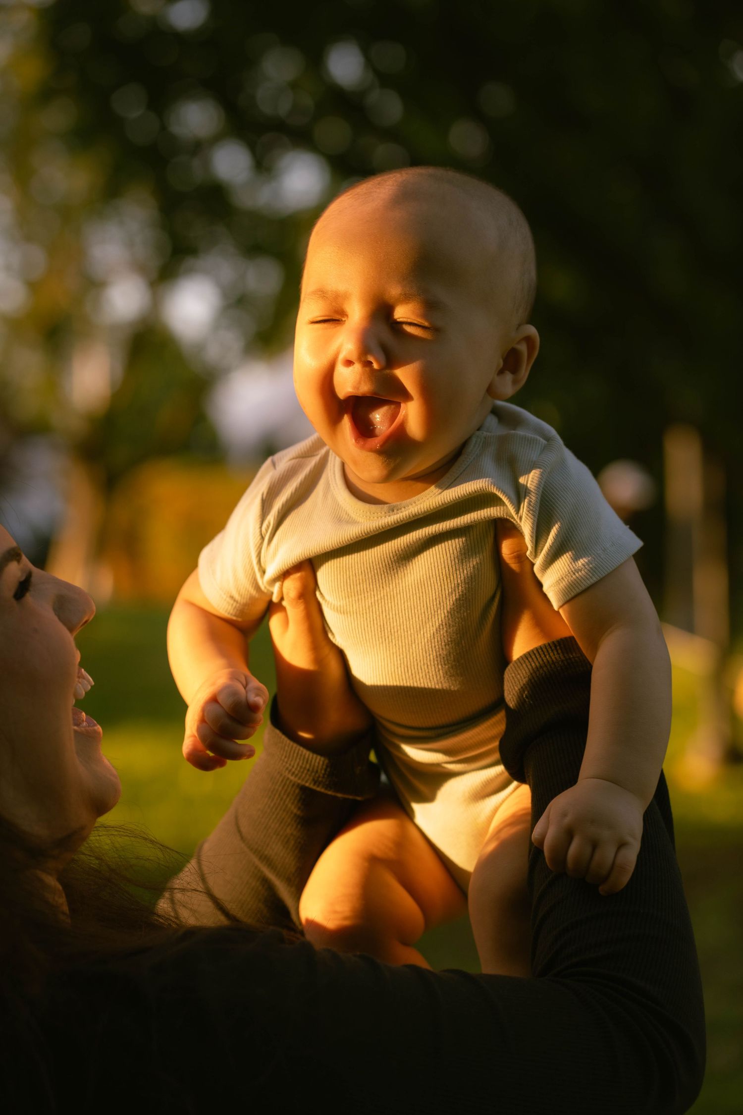 A joyful baby laughs with delight while held up by his mother in warm golden sunset light outdoors.