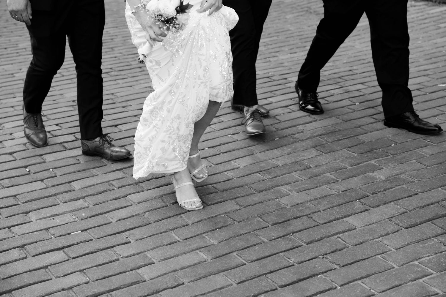 Bride in a lace gown and sandals walks along a brick path beside guests in dark dress shoes in black and white.