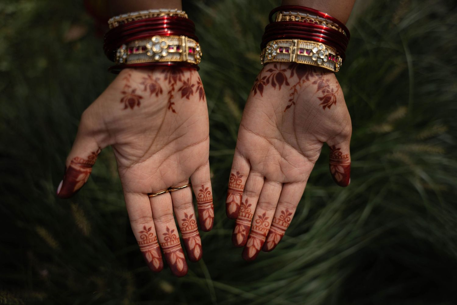 Two open palms decorated with intricate red henna designs, adorned with gold and red bangles, held up against a blurred green background.