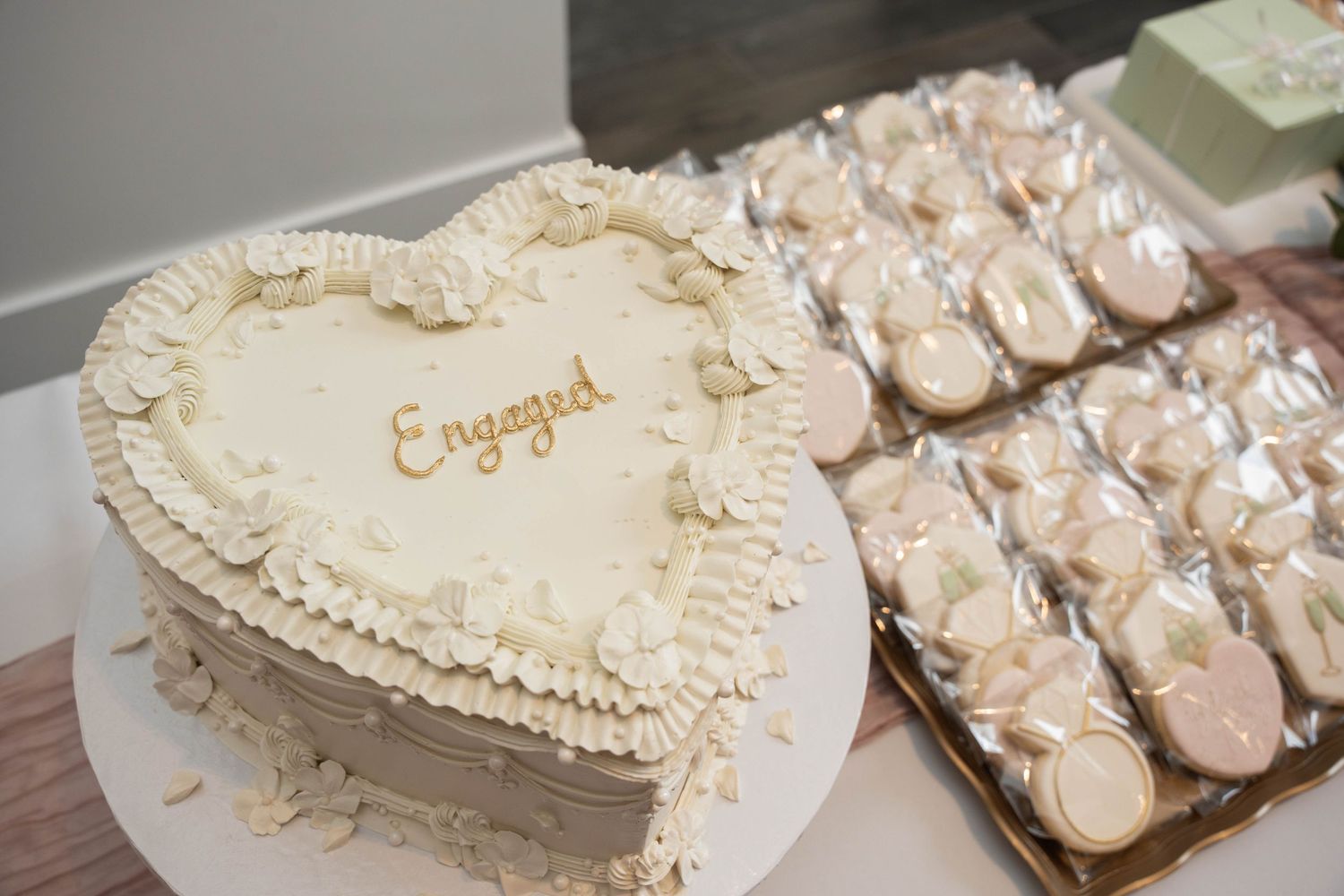 Heart-shaped white engagement cake with the word Engaged piped in gold, next to trays of heart-shaped decorated cookies.