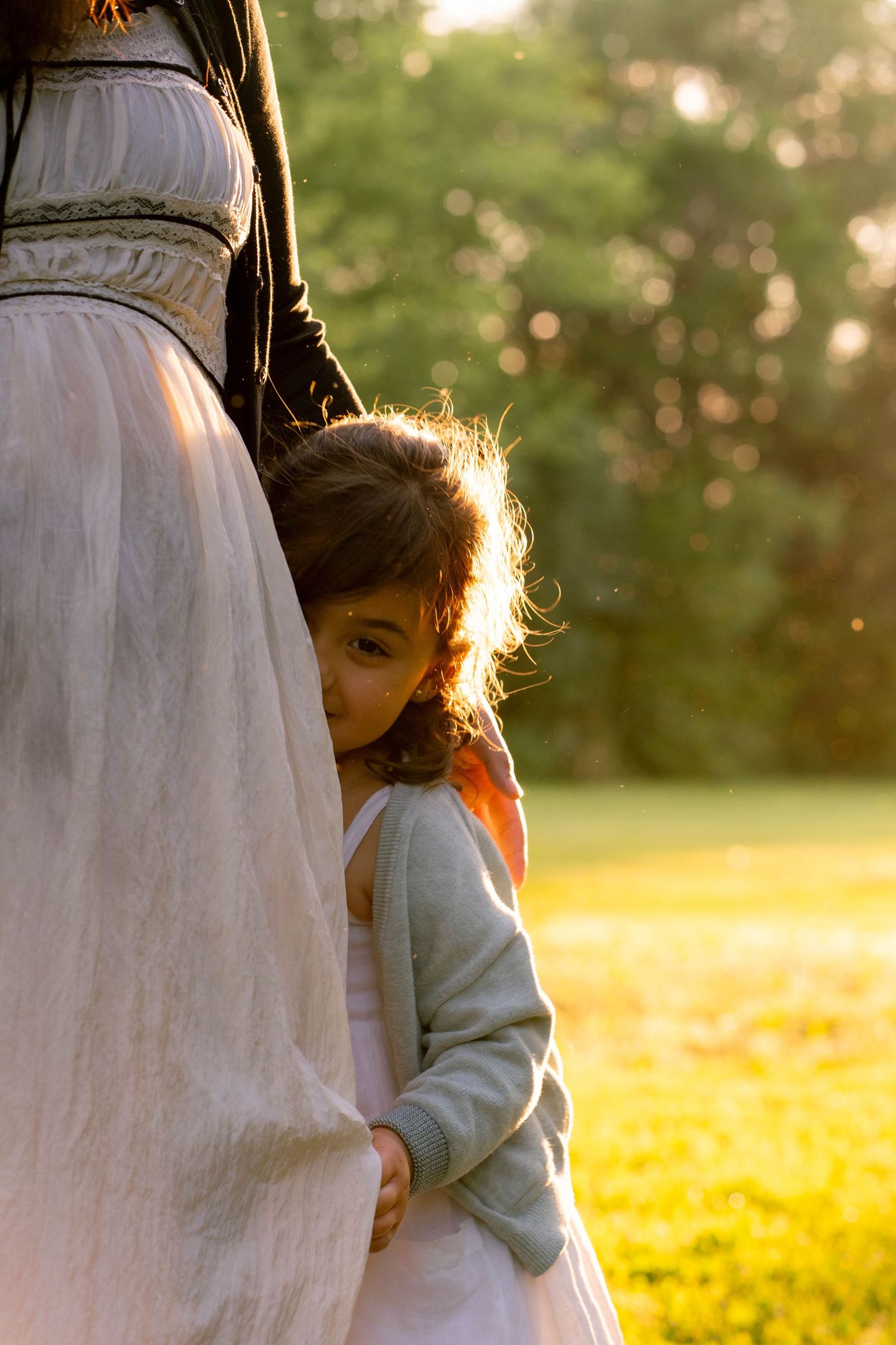 A shy little girl in a gray cardigan peeks from behind her mother's white dress in warm golden evening light.