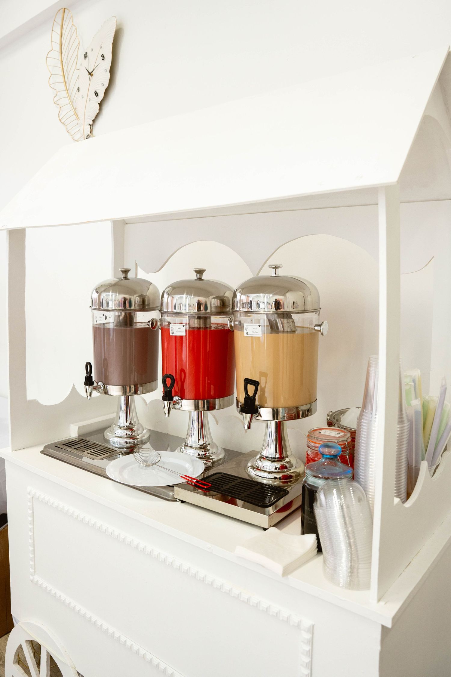 Three chrome beverage dispensers with brown, red and orange drinks sit on a white decorative cart with cups.