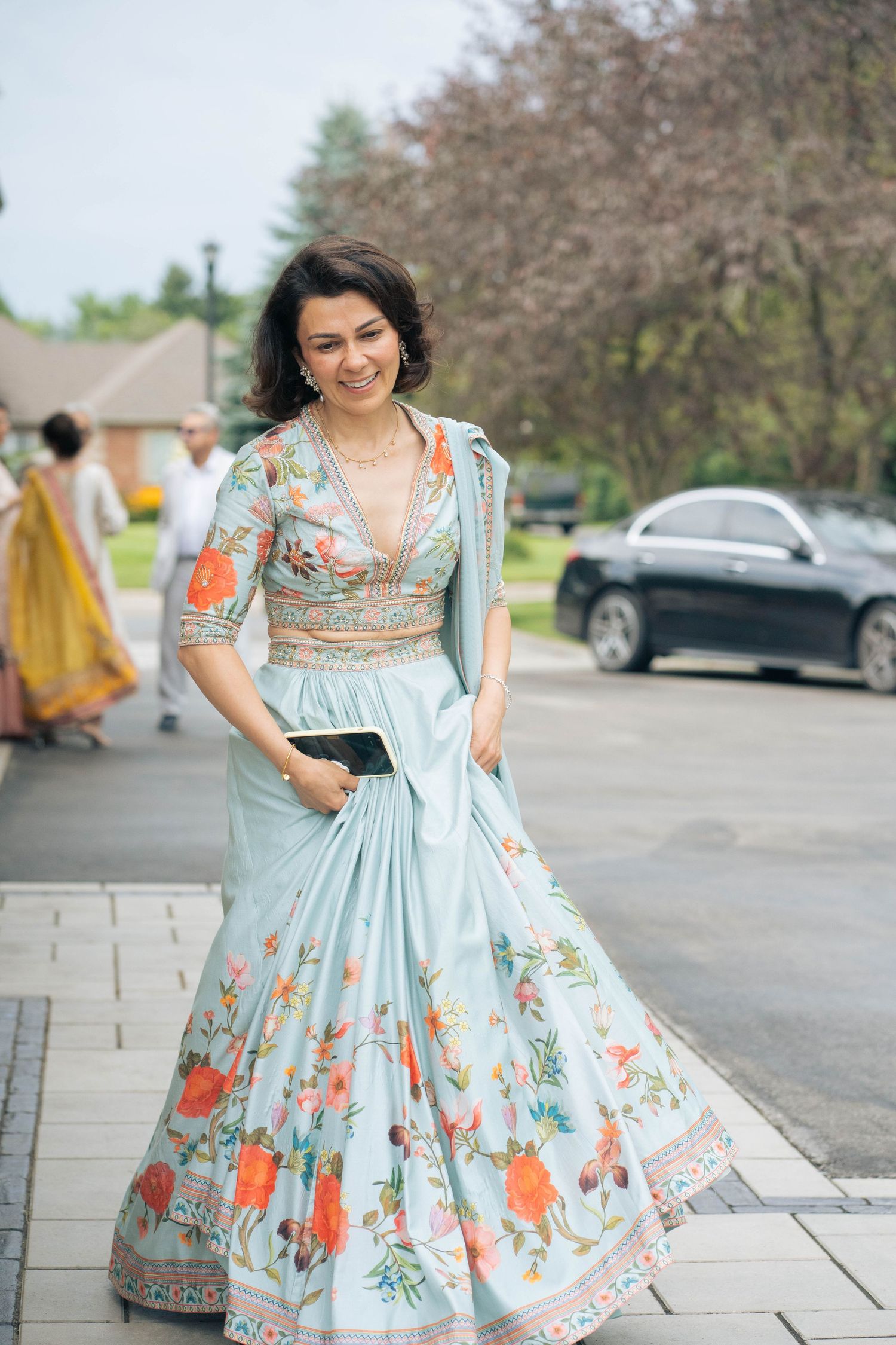 Woman in a light blue floral embroidered lehenga holds a clutch and smiles while standing on a paved walkway outdoors.