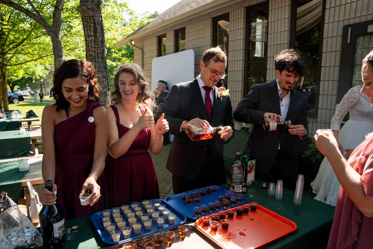 Wedding guests in formal attire serve and enjoy drinks from colorful trays at an outdoor reception under tall trees.