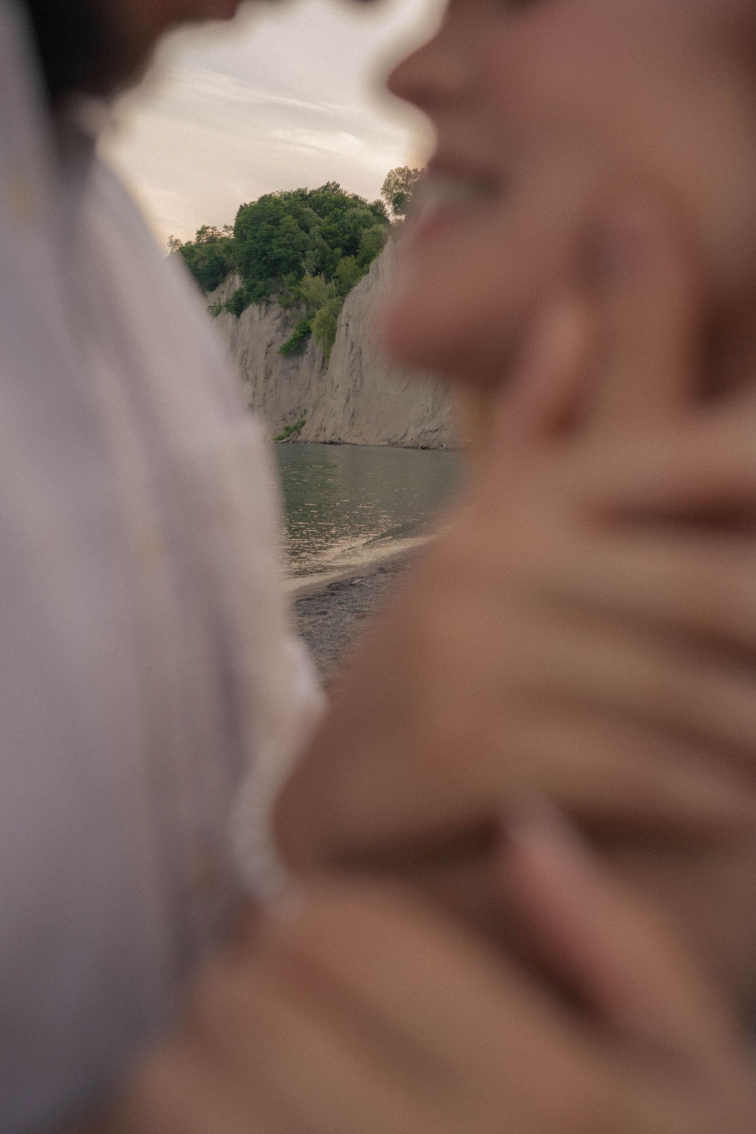 Softly blurred close-up of a couple embracing, with a lake and tree-topped cliff visible in the background.
