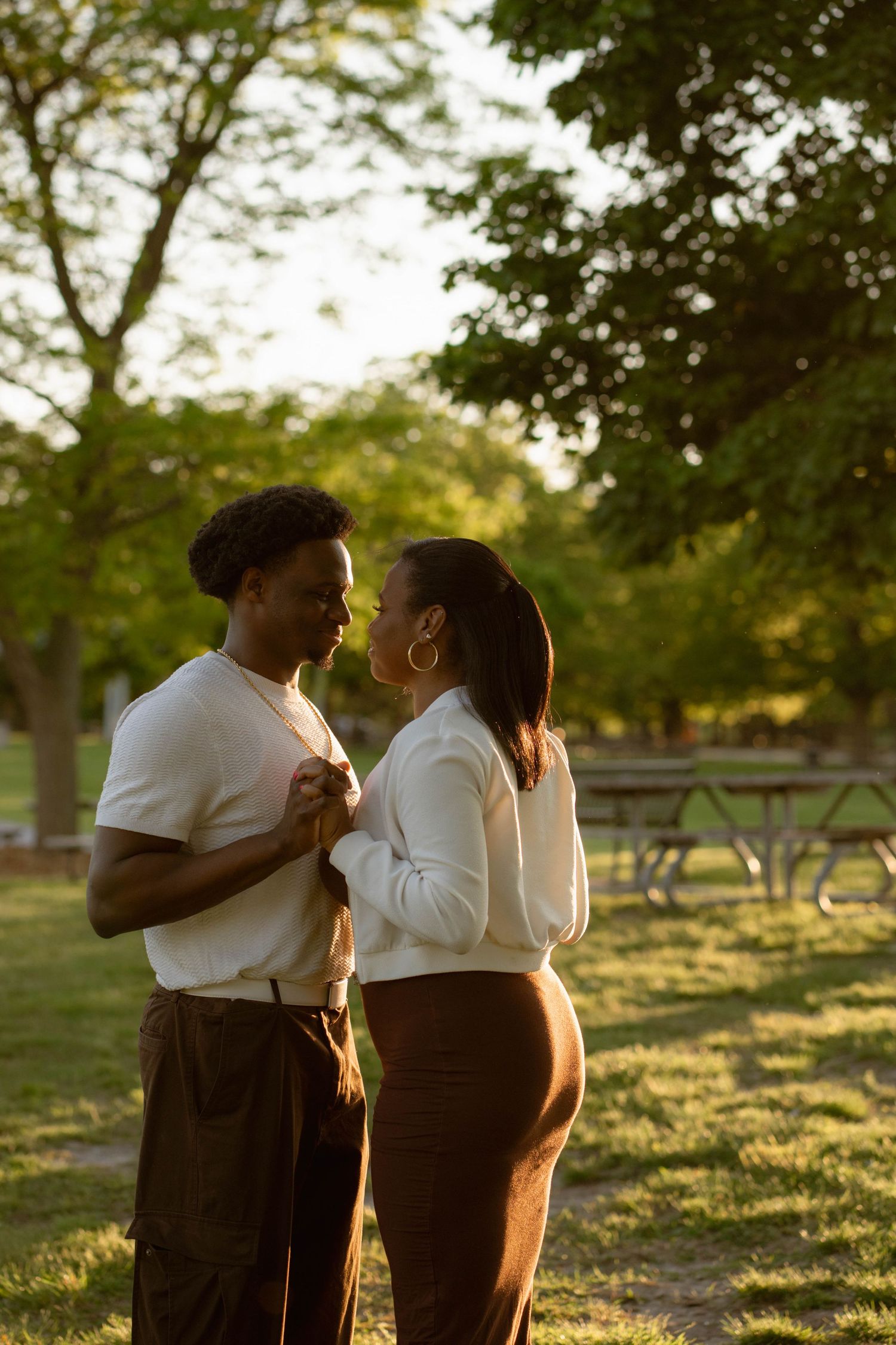 Couple standing face to face and holding hands in a sunlit park, surrounded by trees during a golden hour engagement photo session.