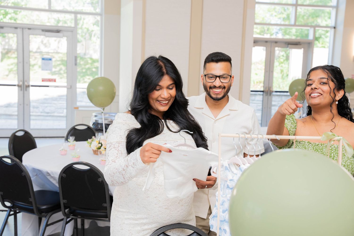 Pregnant woman in a white lace dress opens a card with her husband and a smiling guest at a baby shower with green balloons.