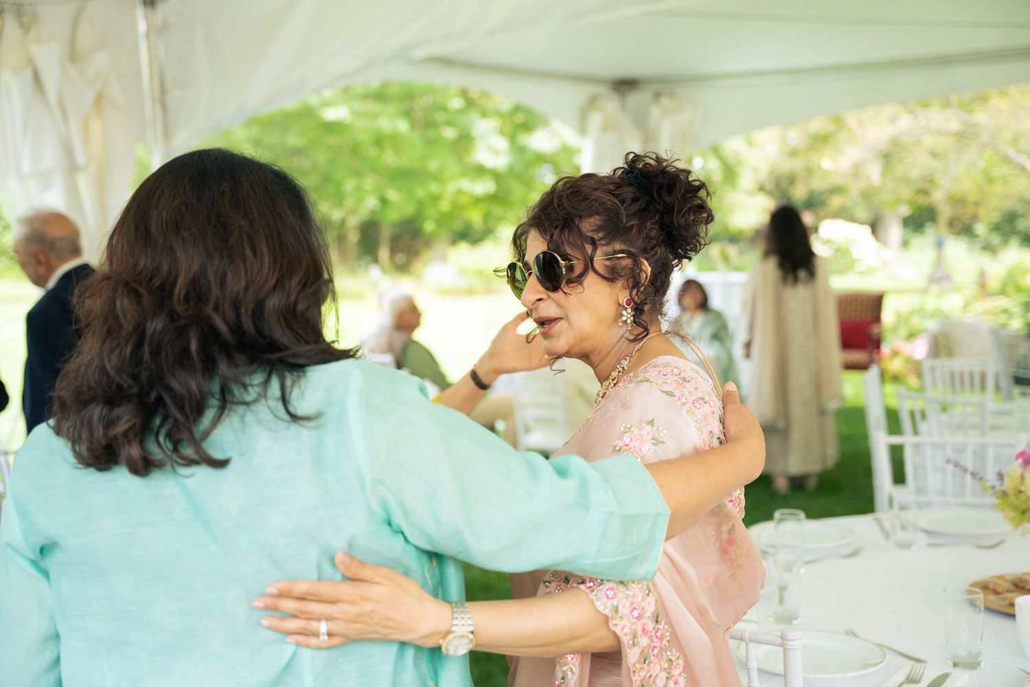 Two women embrace at an outdoor reception table, one in pink with sunglasses and an updo, the other in a teal outfit.
