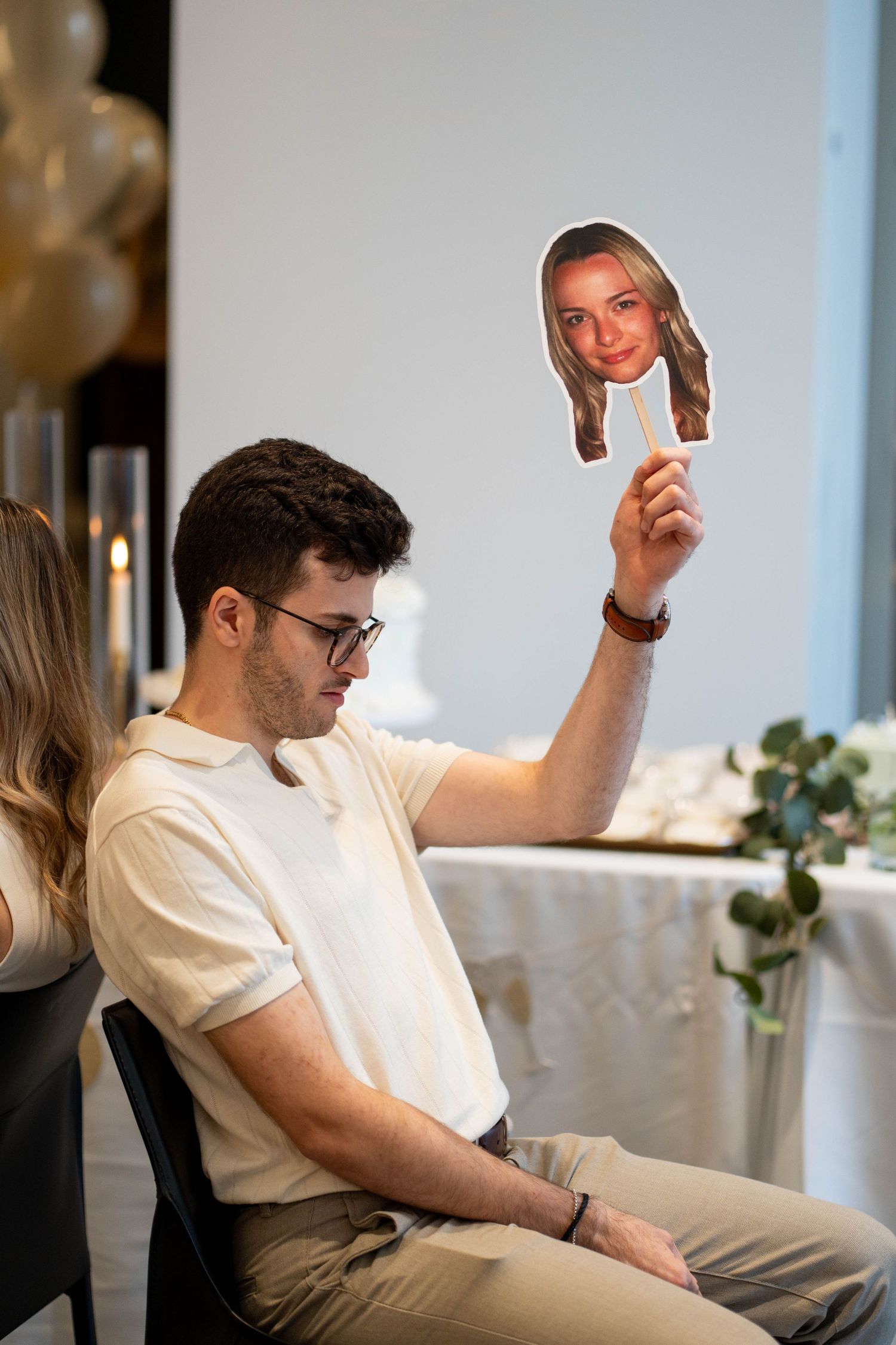 A man in glasses and a cream polo raises a cardboard cutout of a smiling woman's face during an engagement party game.