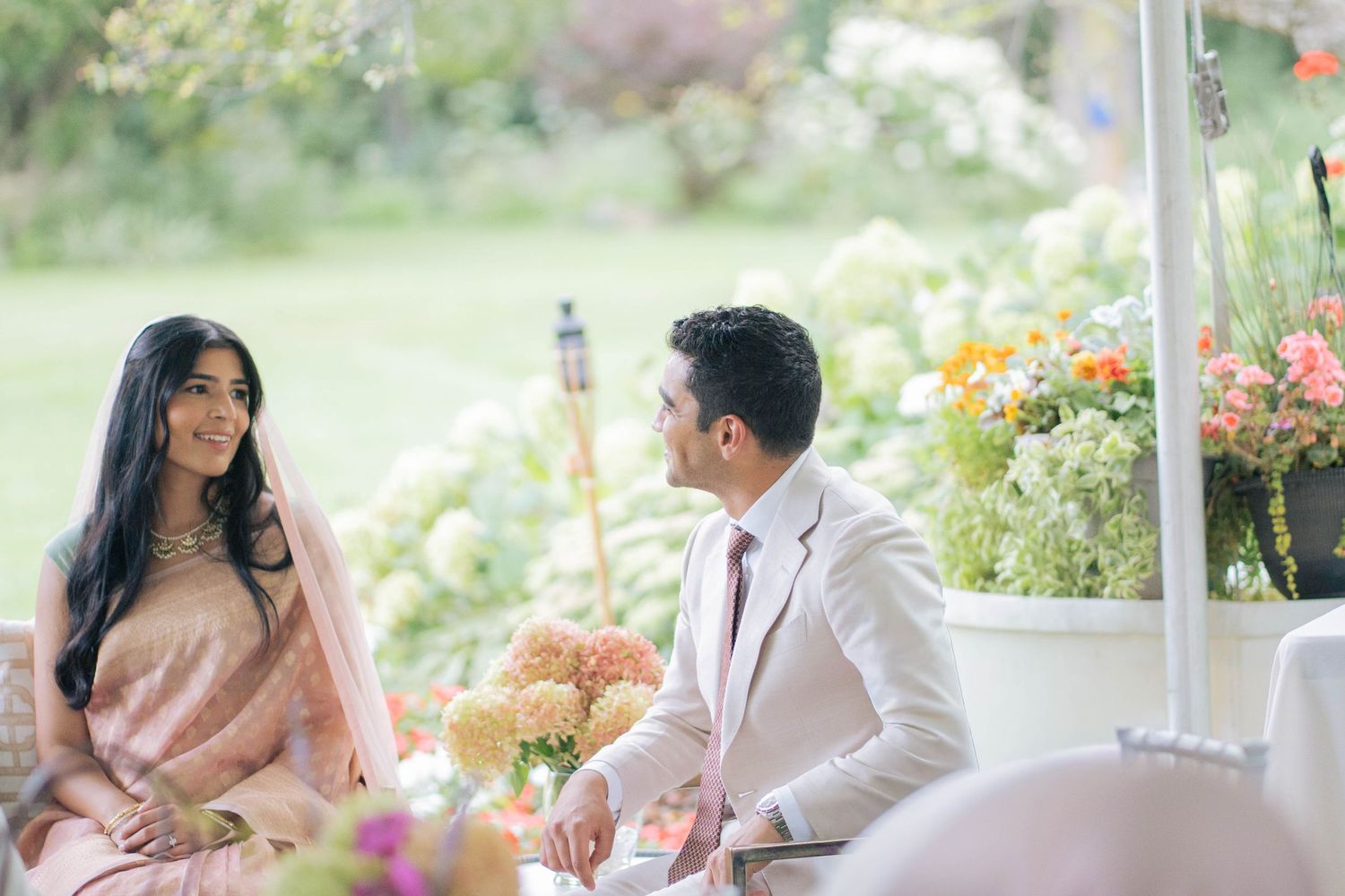 A bride in a peach sari and a groom in a cream suit smile at each other beside pastel flower arrangements in a garden.