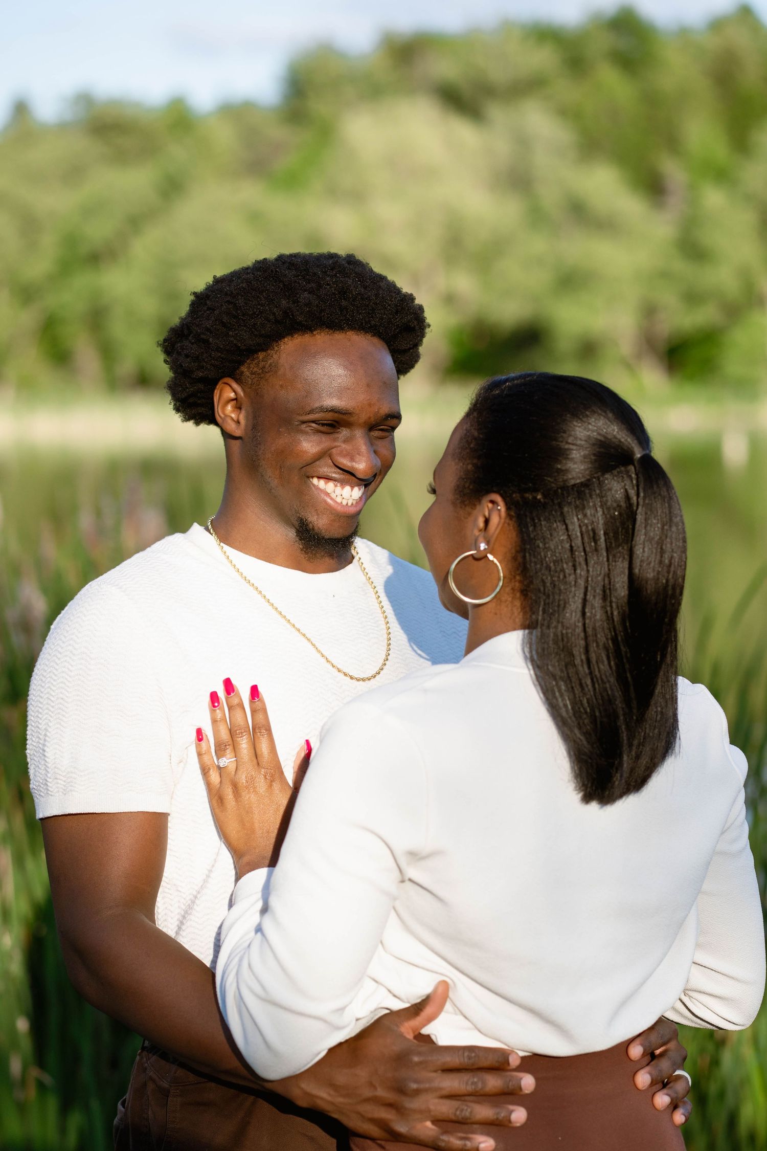 A happy couple embraces in a grassy lakeside park as the woman shows her engagement ring on her partner's chest.