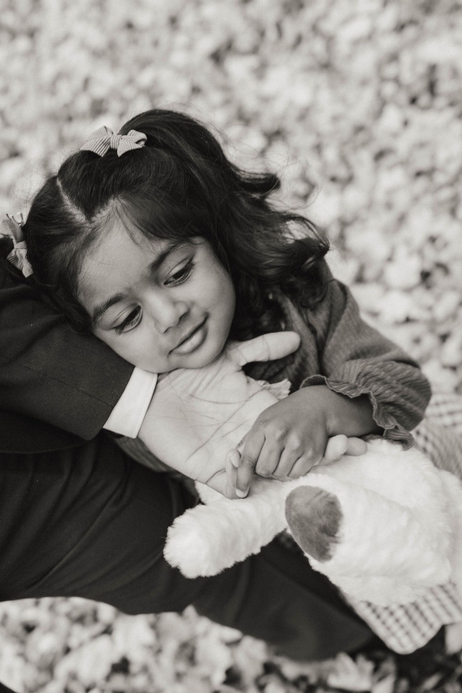 Black and white portrait of a little girl with hair clips resting her head on her father's shoulder while holding a plush lamb.