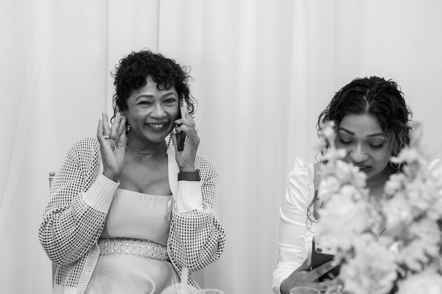 Two women sit near white flowers at a celebration, one smiling while talking on her phone in black and white.
