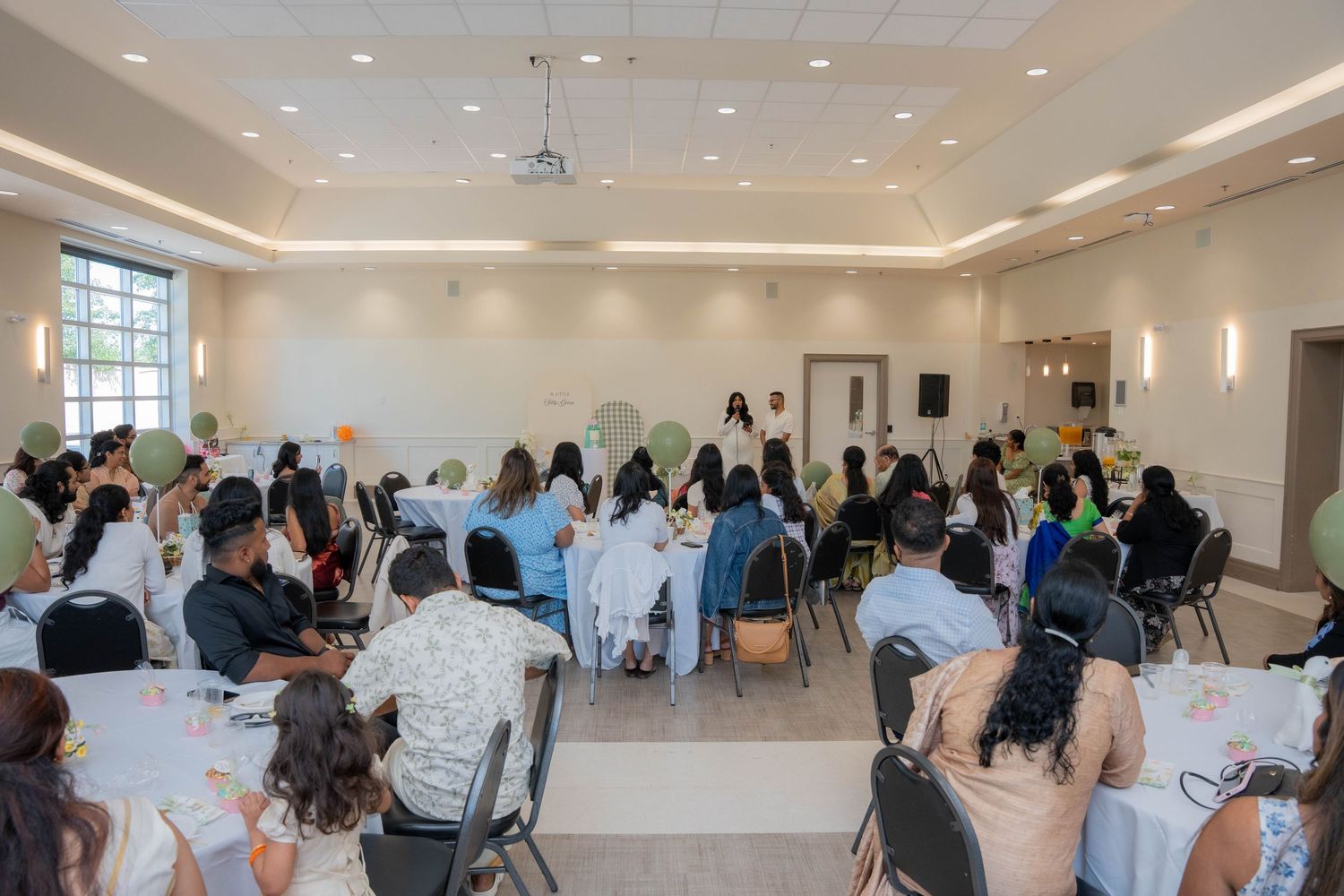 Guests seated at round tables in a bright banquet hall listen to a speaker during a baby shower celebration.