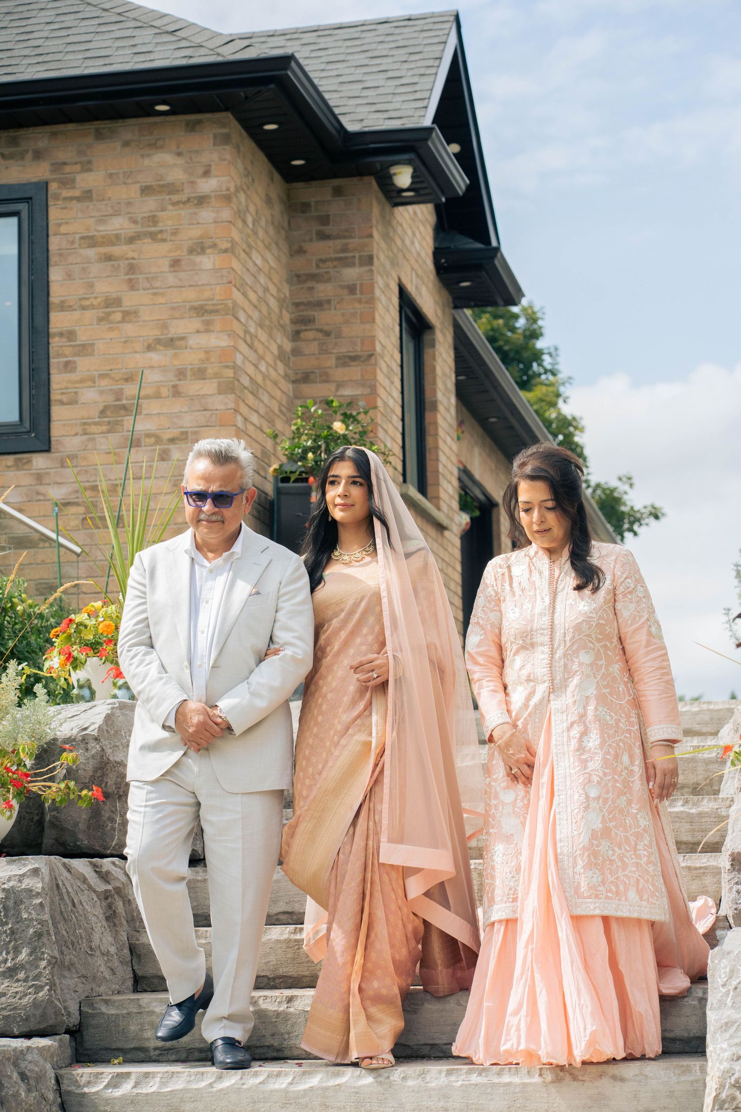 A father in a cream suit walks down stone steps beside a bride in a peach sari and a woman in a pink outfit at a brick home.