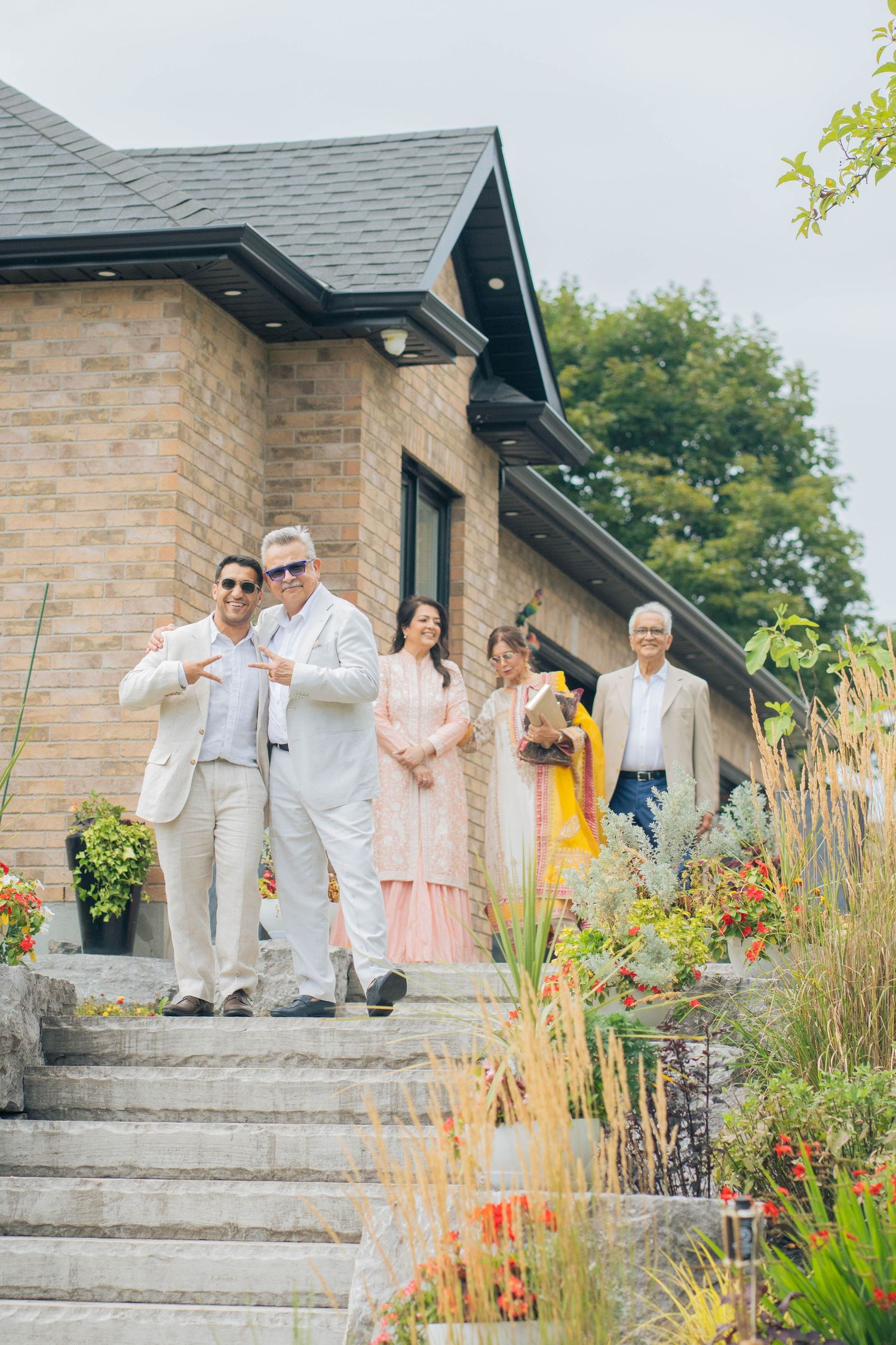 Family and guests in light formal and traditional outfits pose together on stone steps outside a brick house with gardens.