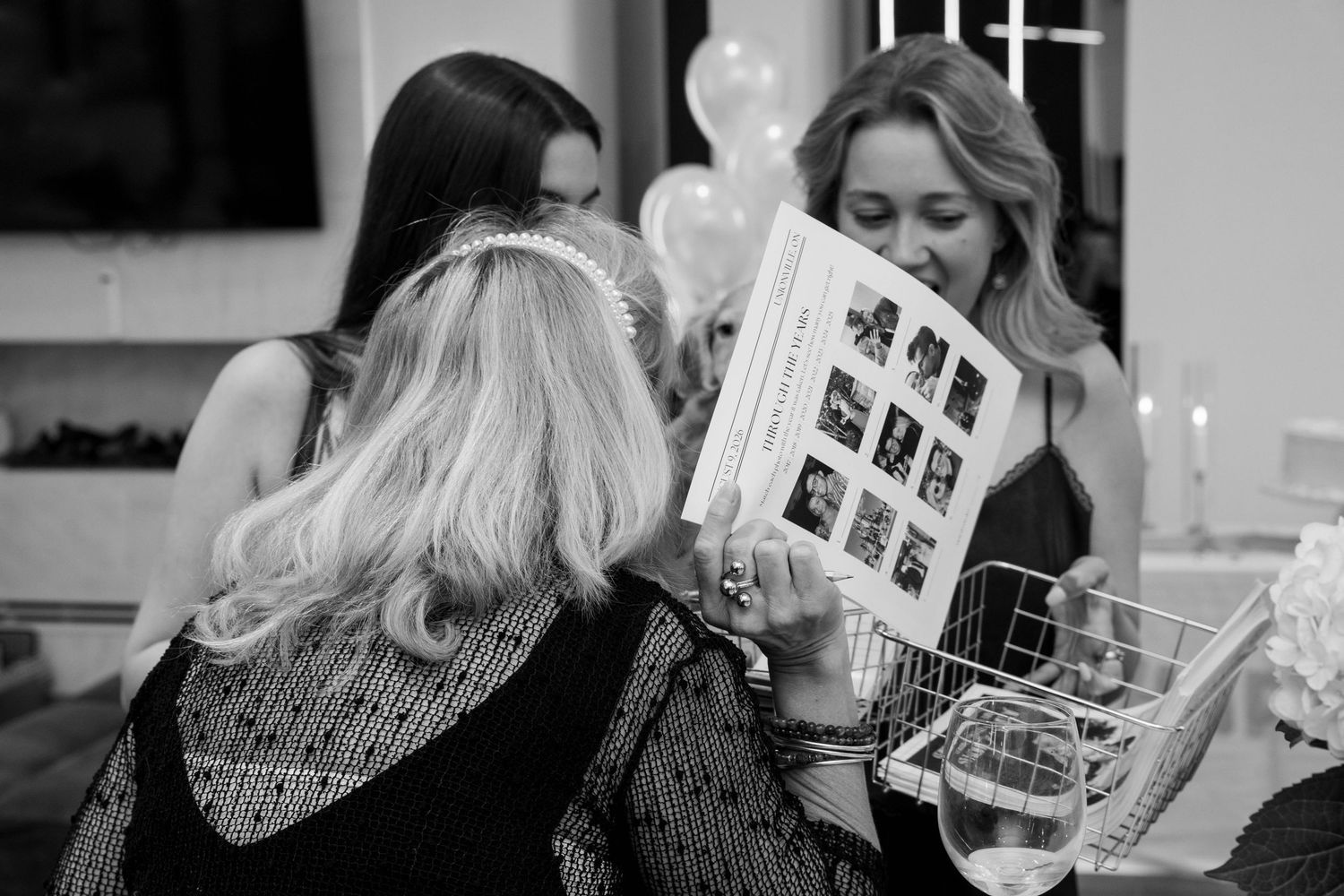Guests in a black and white photo look at a printed trivia sheet with a grid of photos during an engagement party.
