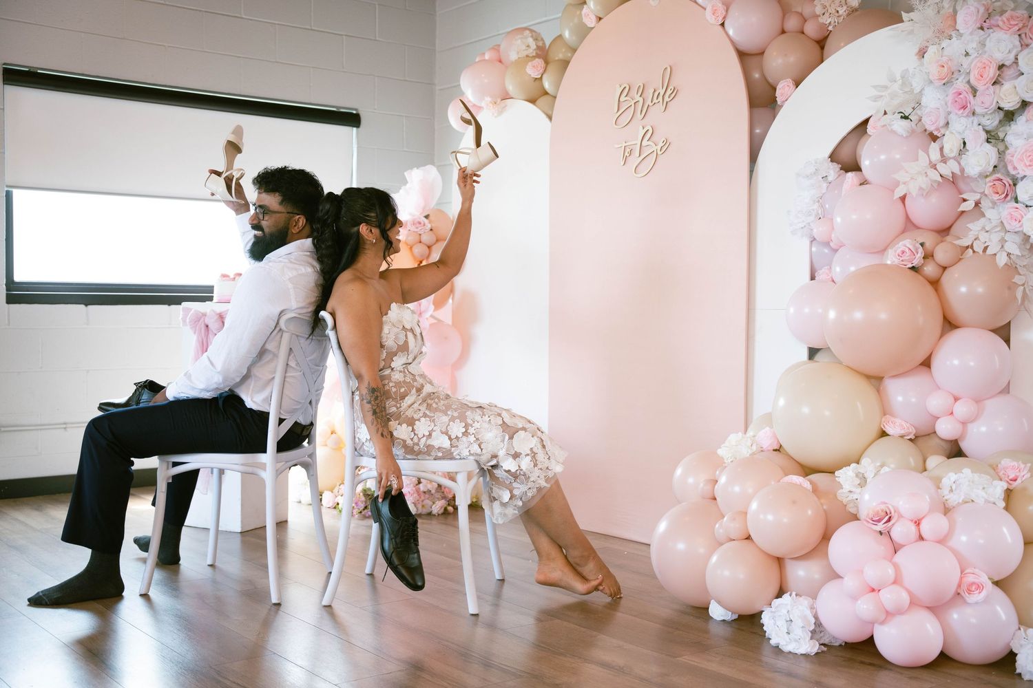 Couple seated back to back raise shoes during a bridal shower game beside a pink balloon garland and Bride to Be backdrop.