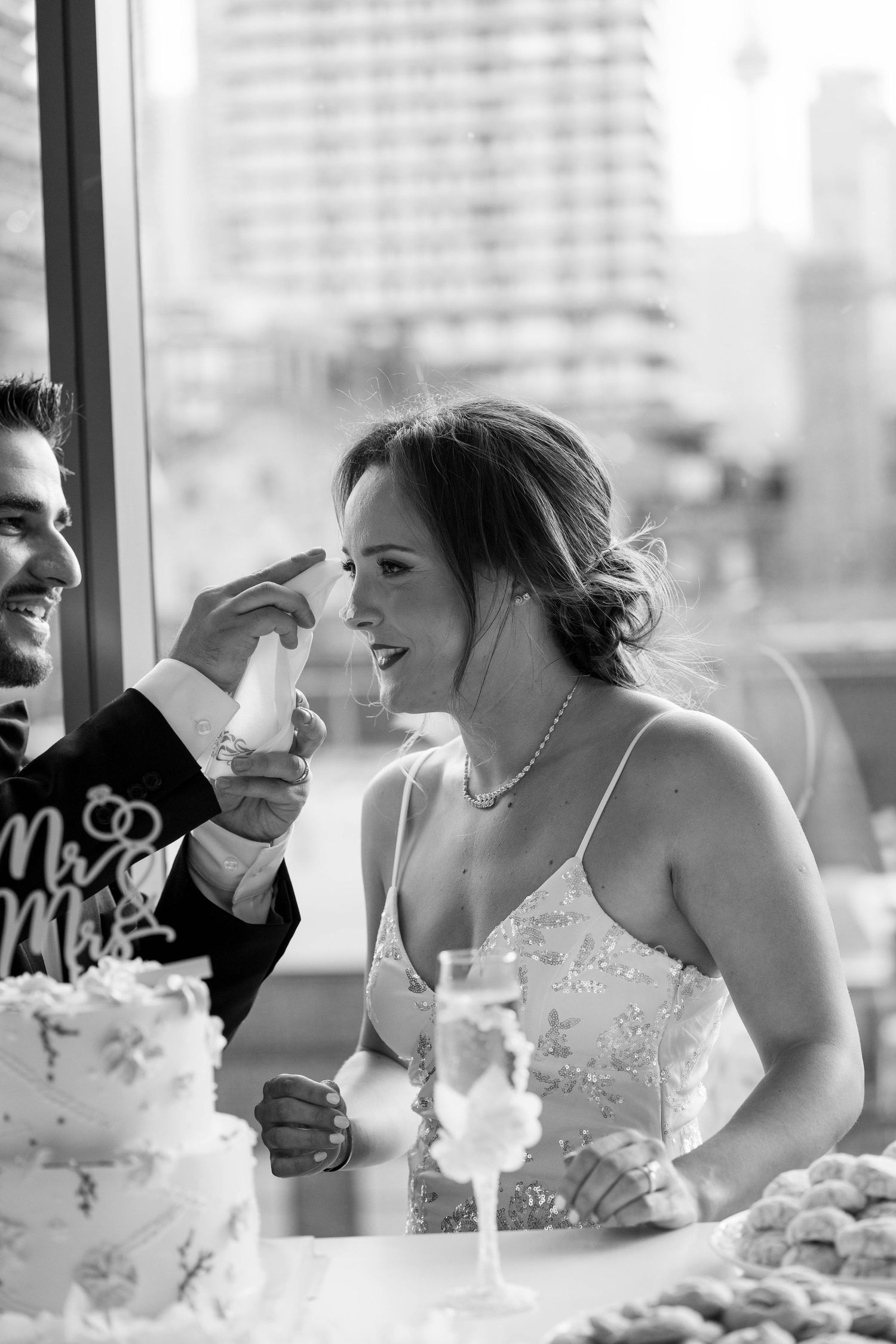 Black and white photo of a groom gently touching his bride's face as she smiles, with a Mr and Mrs cake and champagne nearby.
