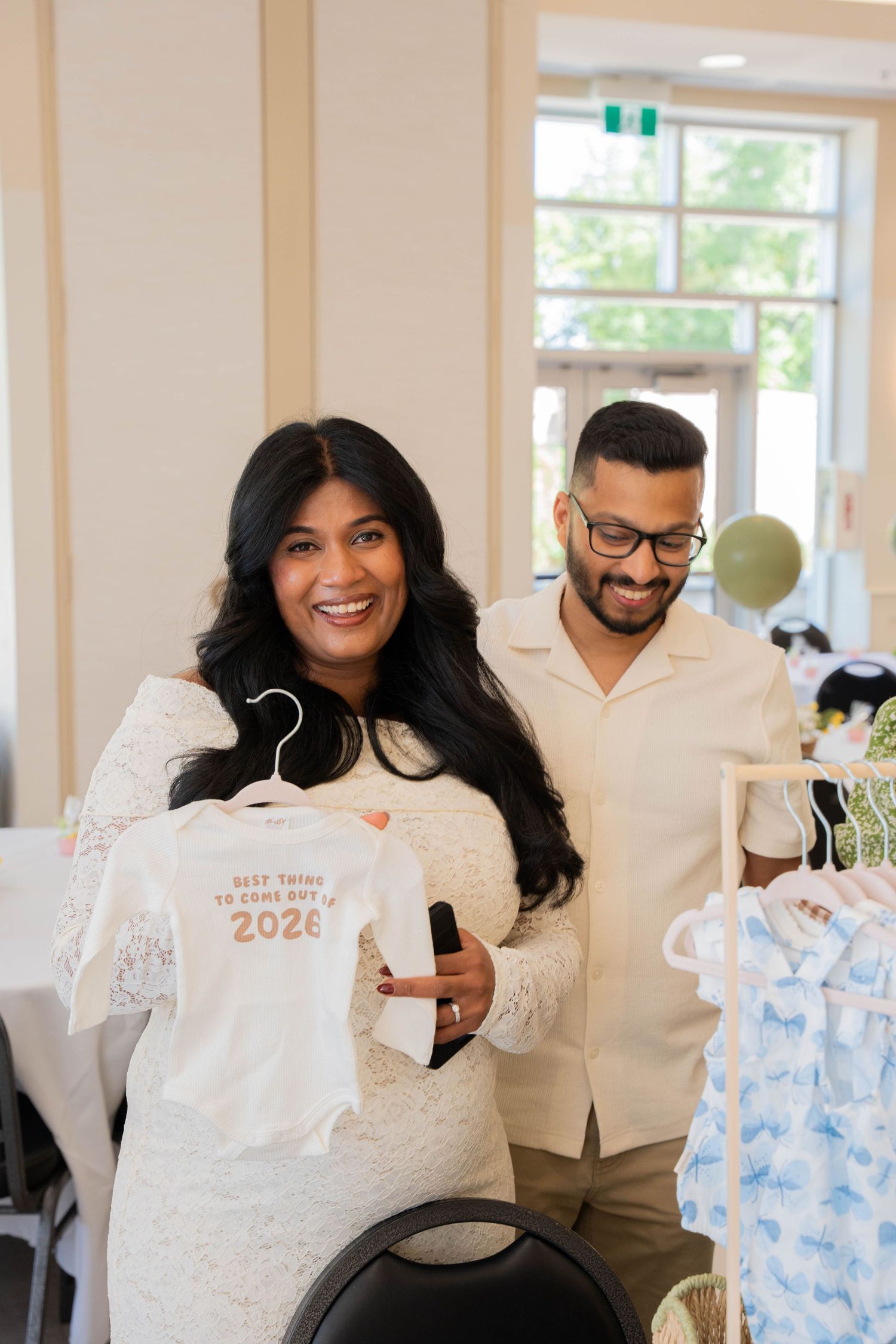 Smiling expectant mother holds a white baby onesie reading 'Best things to come out in 2026' beside her bearded husband in glasses.