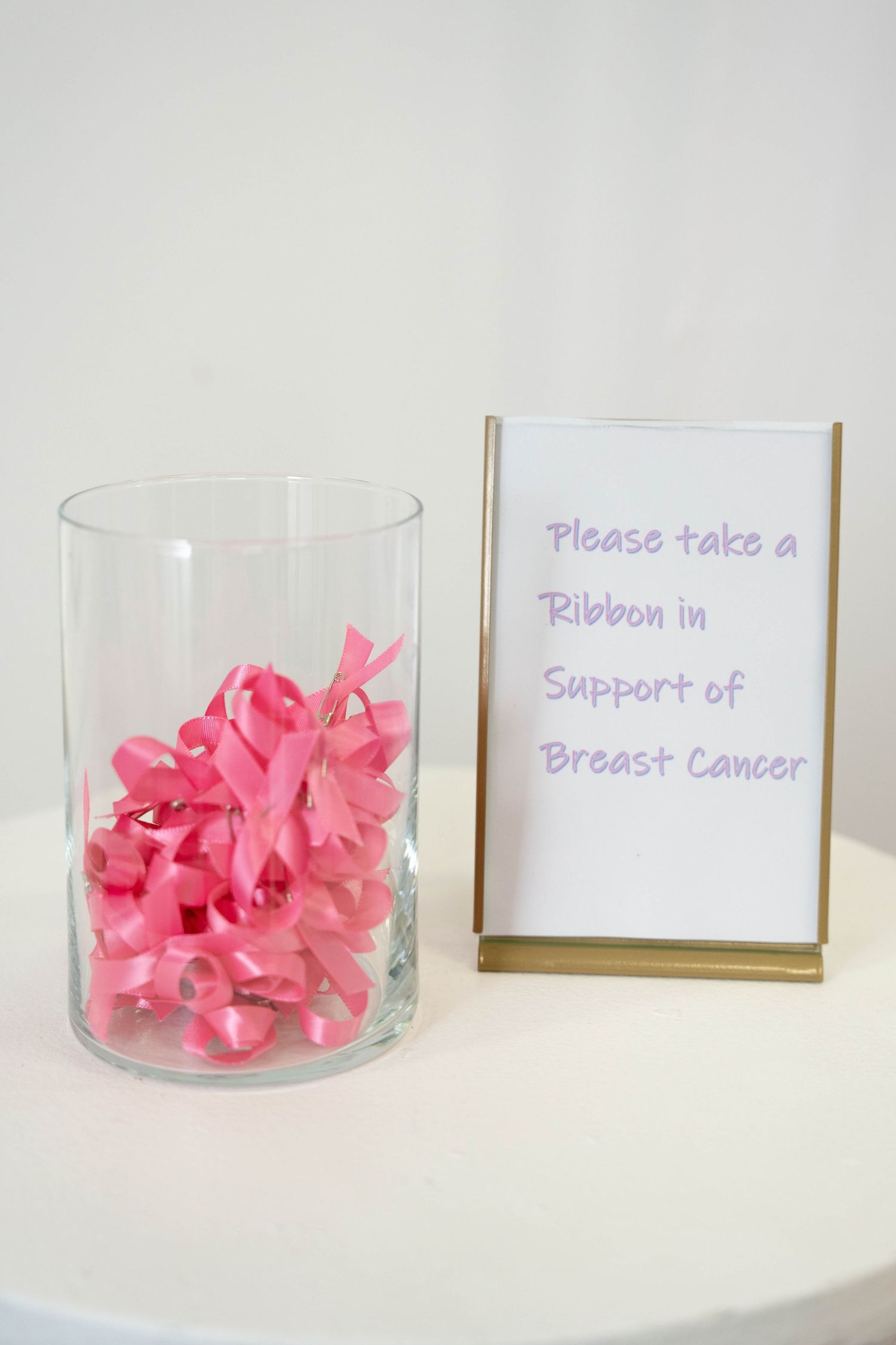 Glass vase of pink ribbons beside a gold-framed sign inviting guests to take a ribbon in support of breast cancer.