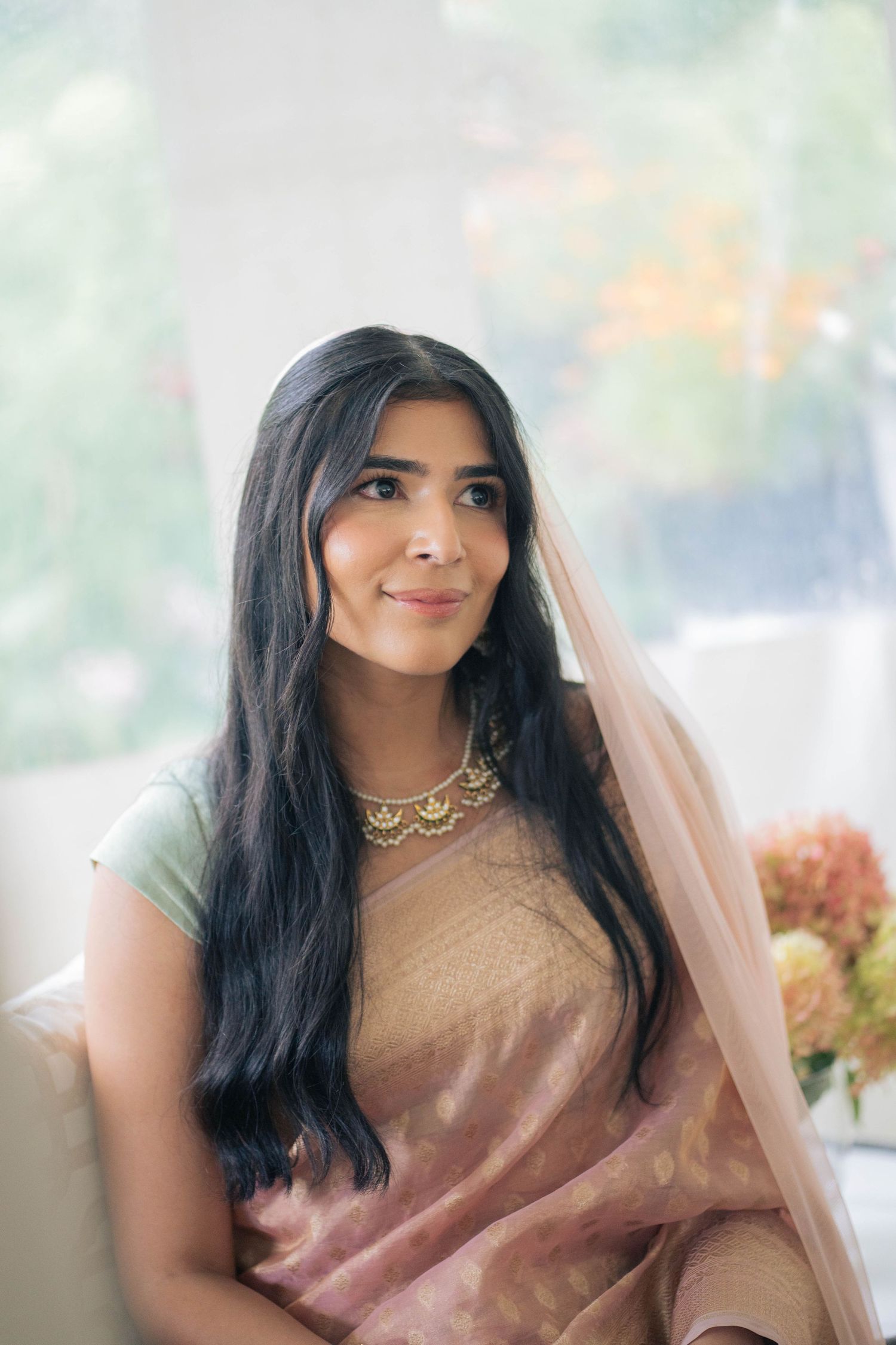Portrait of a bride with long dark hair wearing a peach sari, gold statement necklace and sheer veil, smiling softly beside peach flowers.