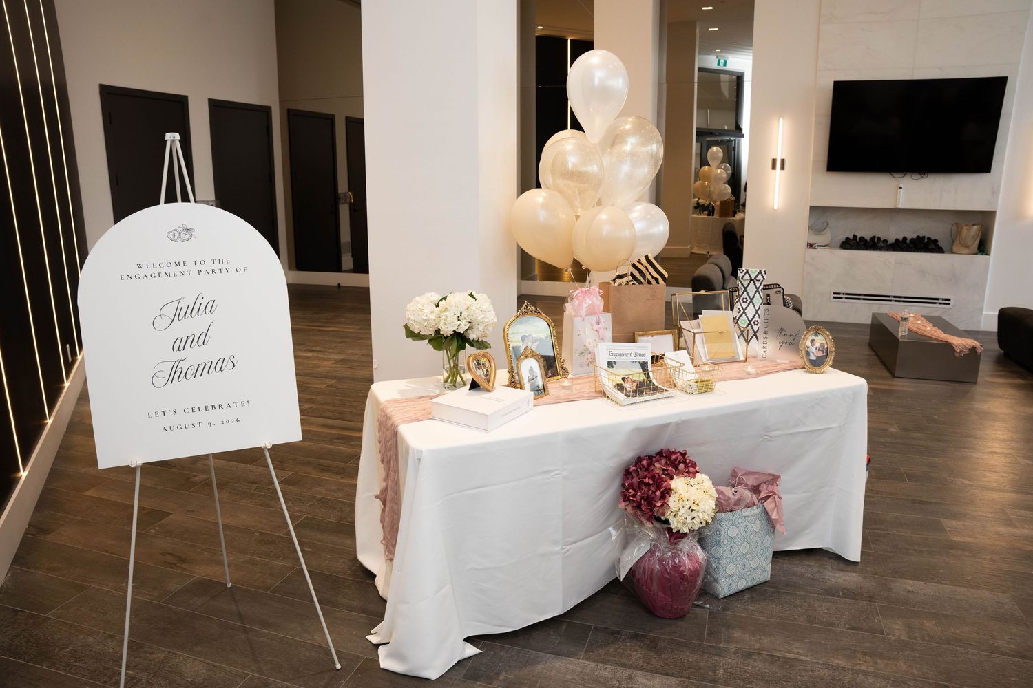 White-draped welcome table with an engagement sign on an easel, floral arrangements, framed photos, and cream balloons.