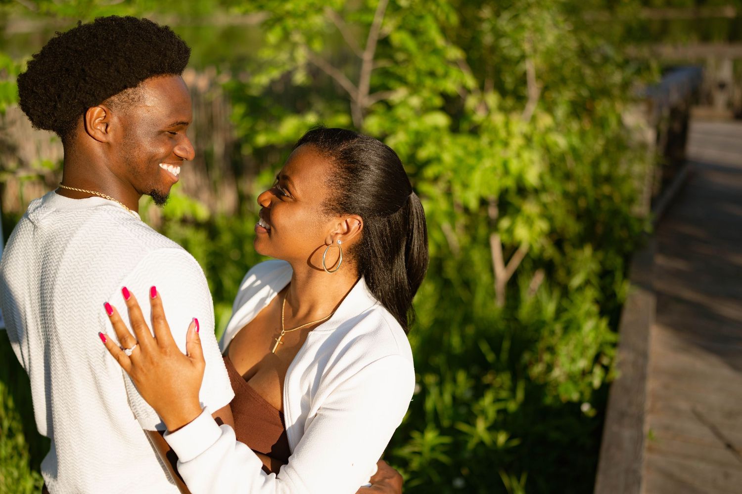 Smiling woman showing her engagement ring as she embraces her partner outdoors among lush green foliage in warm sunlight.
