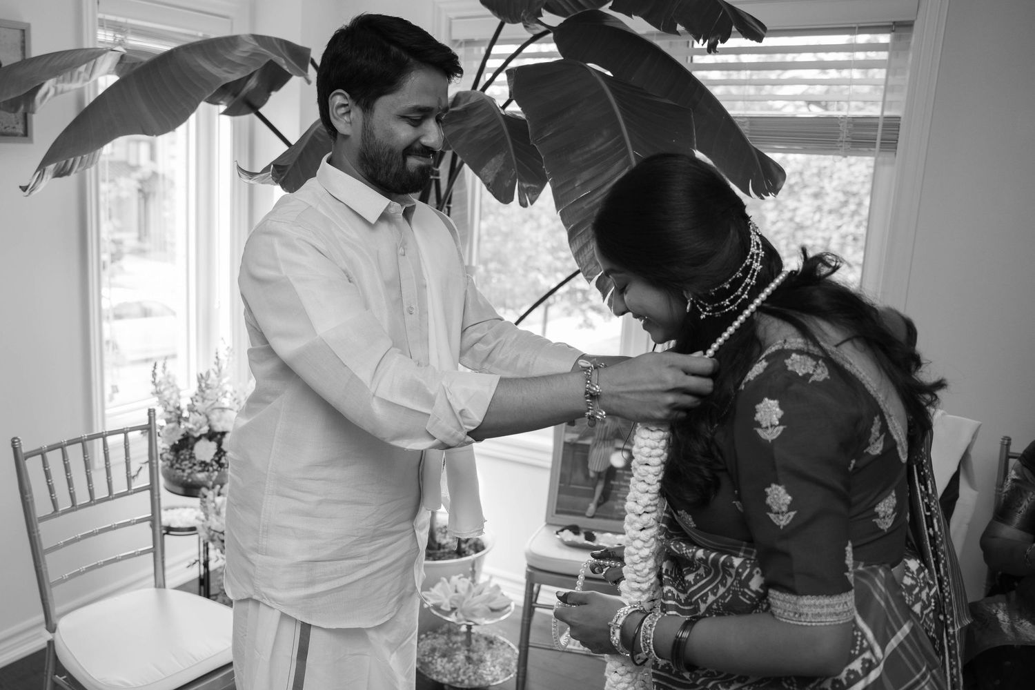 Black and white photo of a groom in a dhoti placing a flower garland around a bride in a patterned saree indoors.