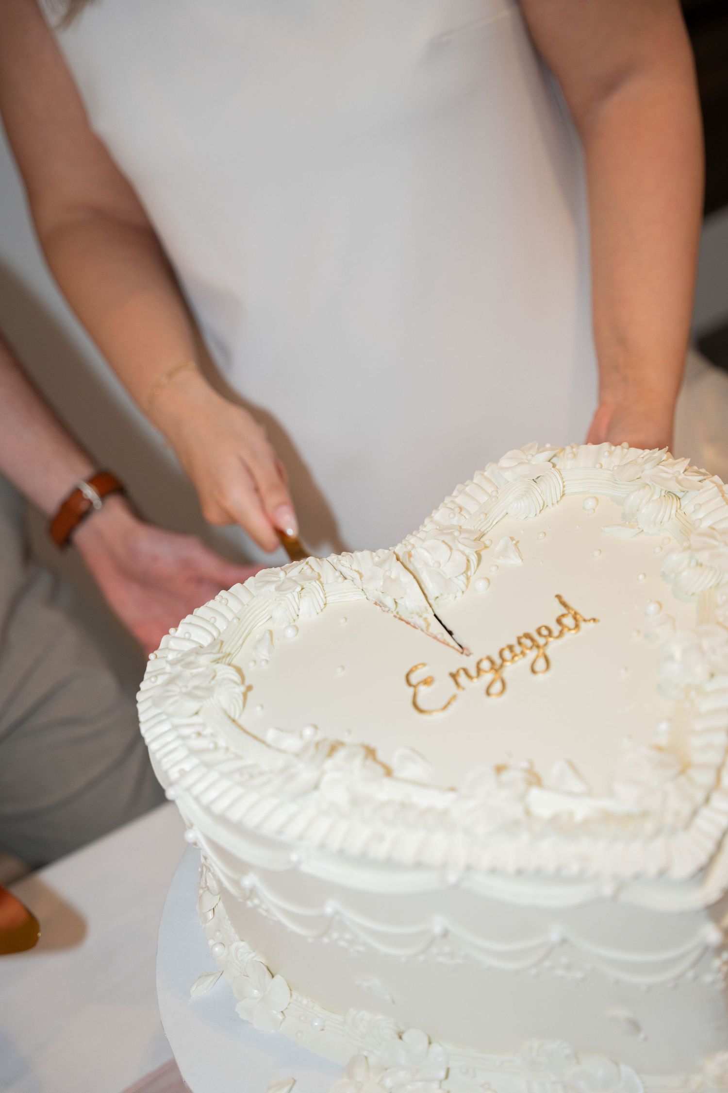 A woman in a white dress cuts into a heart-shaped white engagement cake with gold Engaged lettering and piped frosting borders.