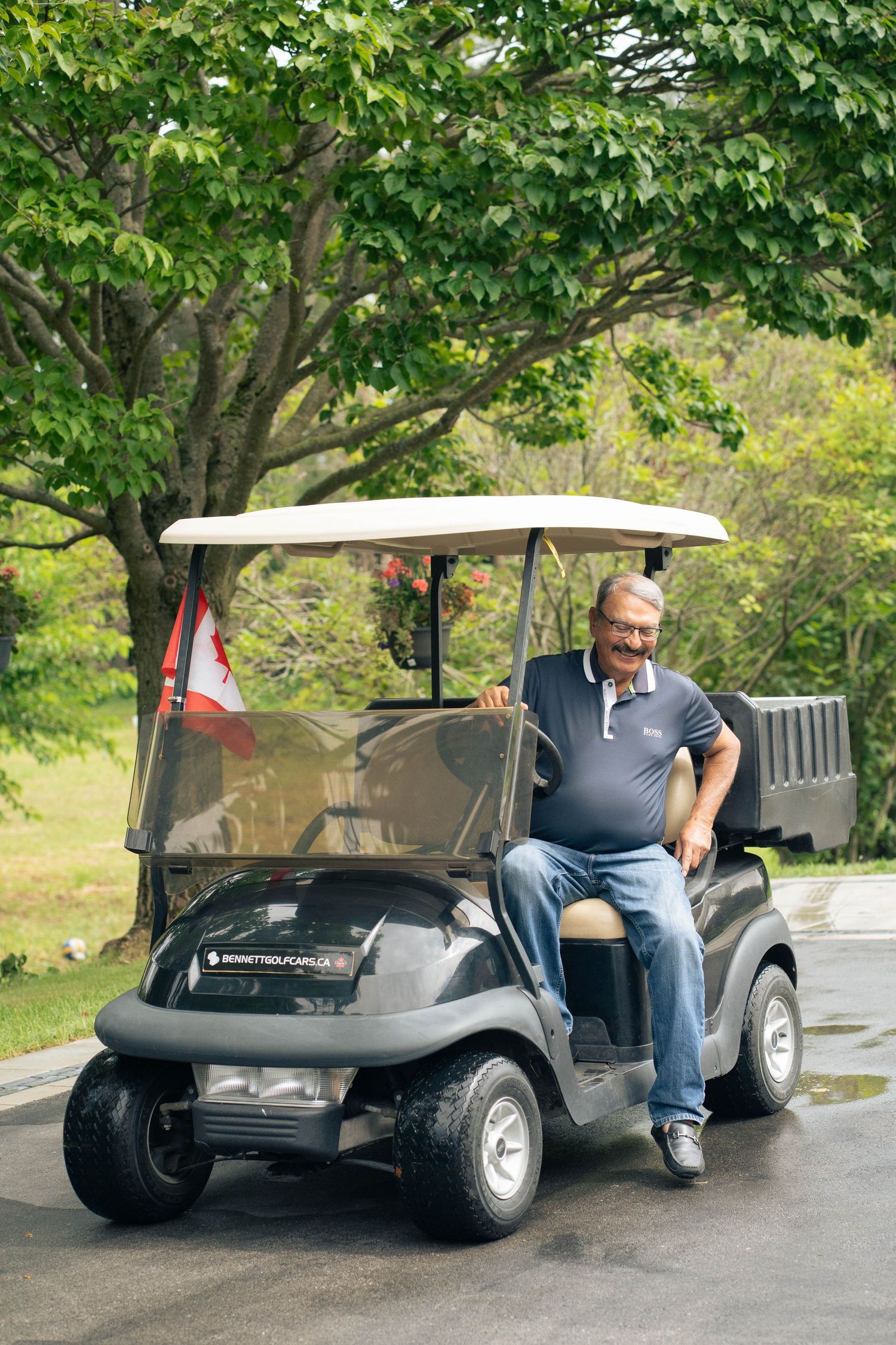 Smiling man in a navy polo sits in a black golf cart parked on a driveway beneath leafy green trees.