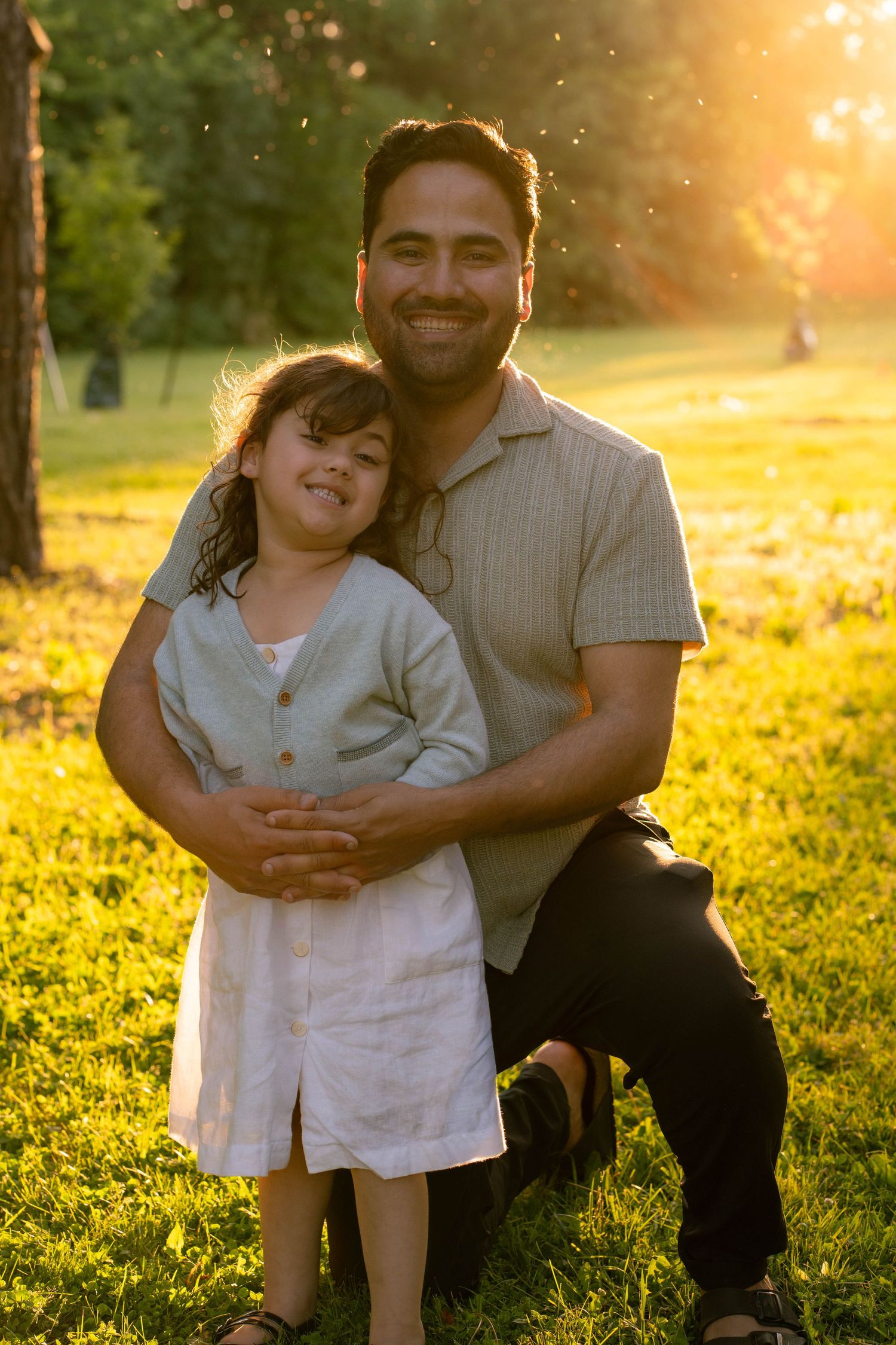 A smiling father kneels in a sunlit grassy field, hugging his happy young daughter in a white linen dress.