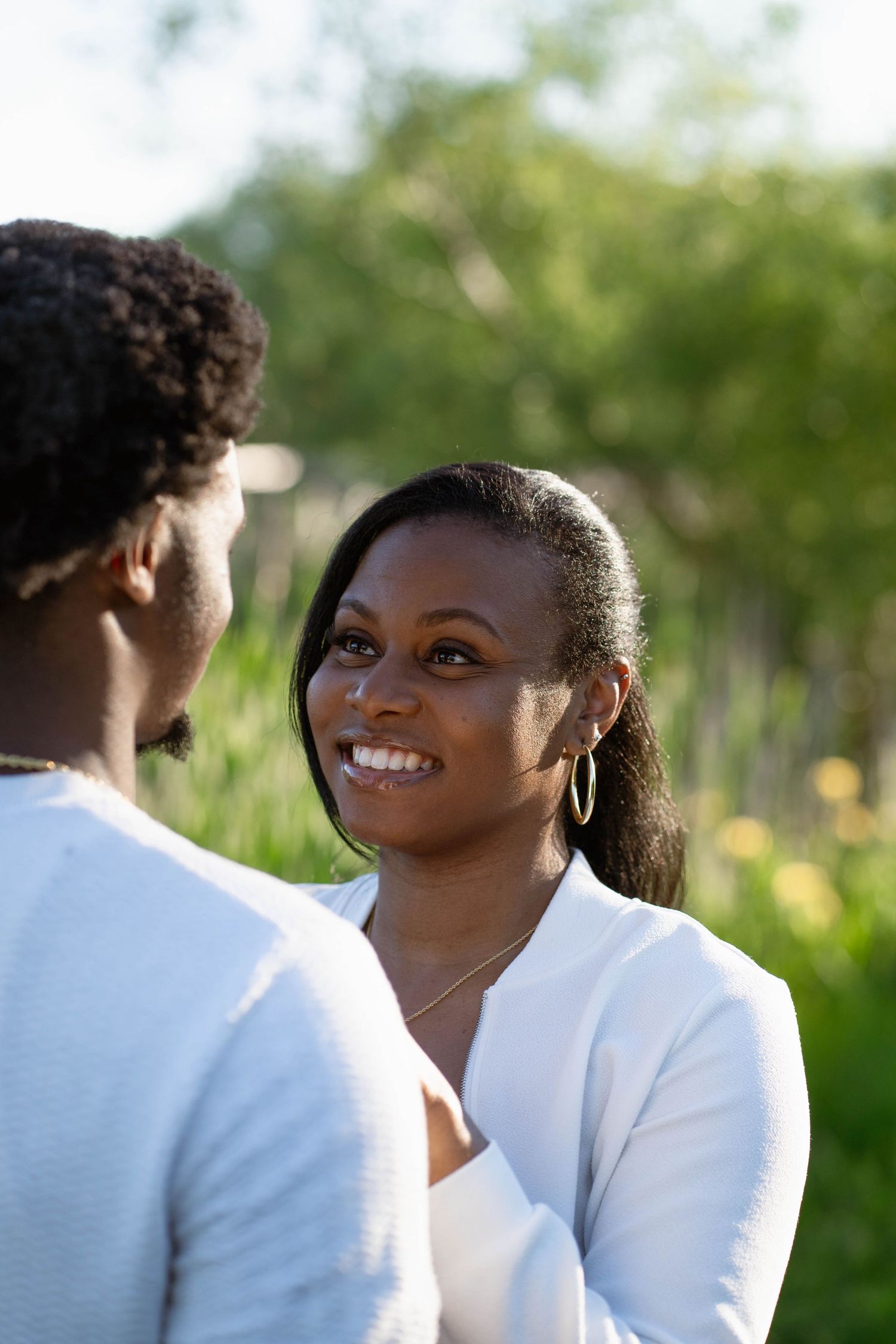 A smiling woman in a white jacket and hoop earrings looks lovingly at her fiancé in a sunlit green field.