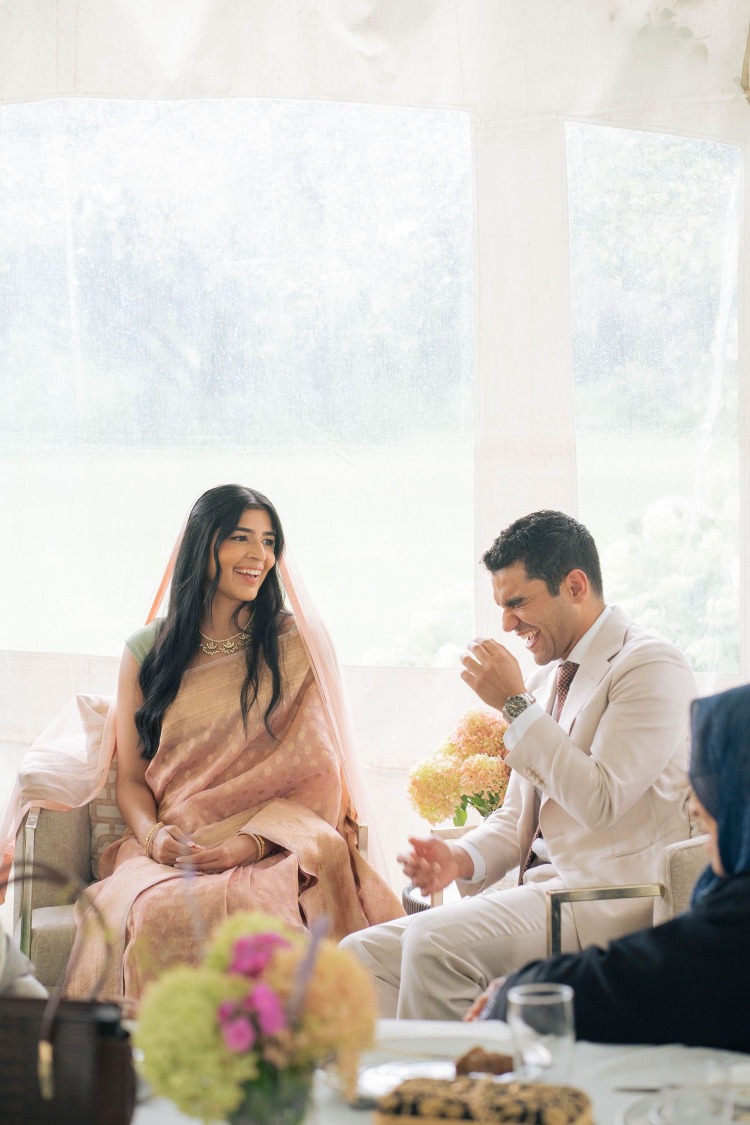 A smiling bride in a peach embroidered outfit and veil laughs with a groom in a cream suit at a bright tent wedding with floral centerpieces