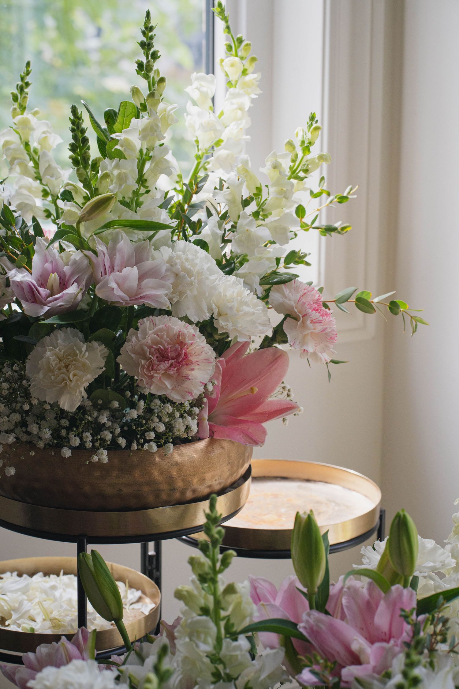 Elegant wedding floral arrangement of white and pink carnations, lilies, and baby's breath in a gold vase near a sunlit window.