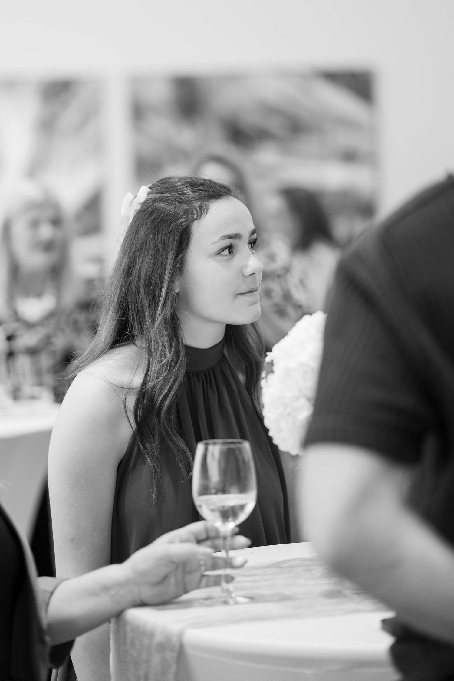 Black and white photo of a young woman with long hair holding a wine glass at a reception table, gazing off to the side.