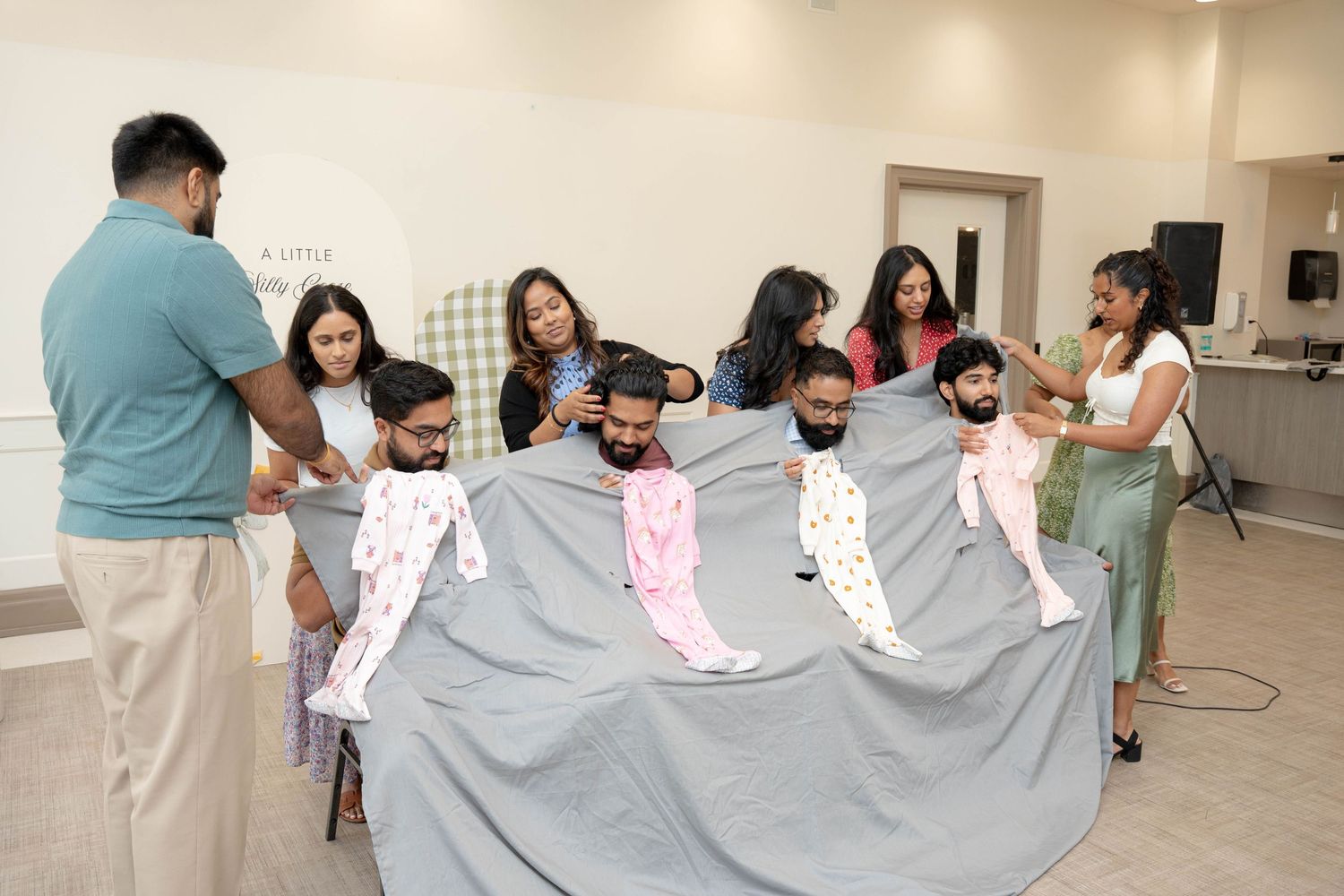 Guests gather around a large gray sheet while several men wear pink baby onesies during a playful baby shower game.