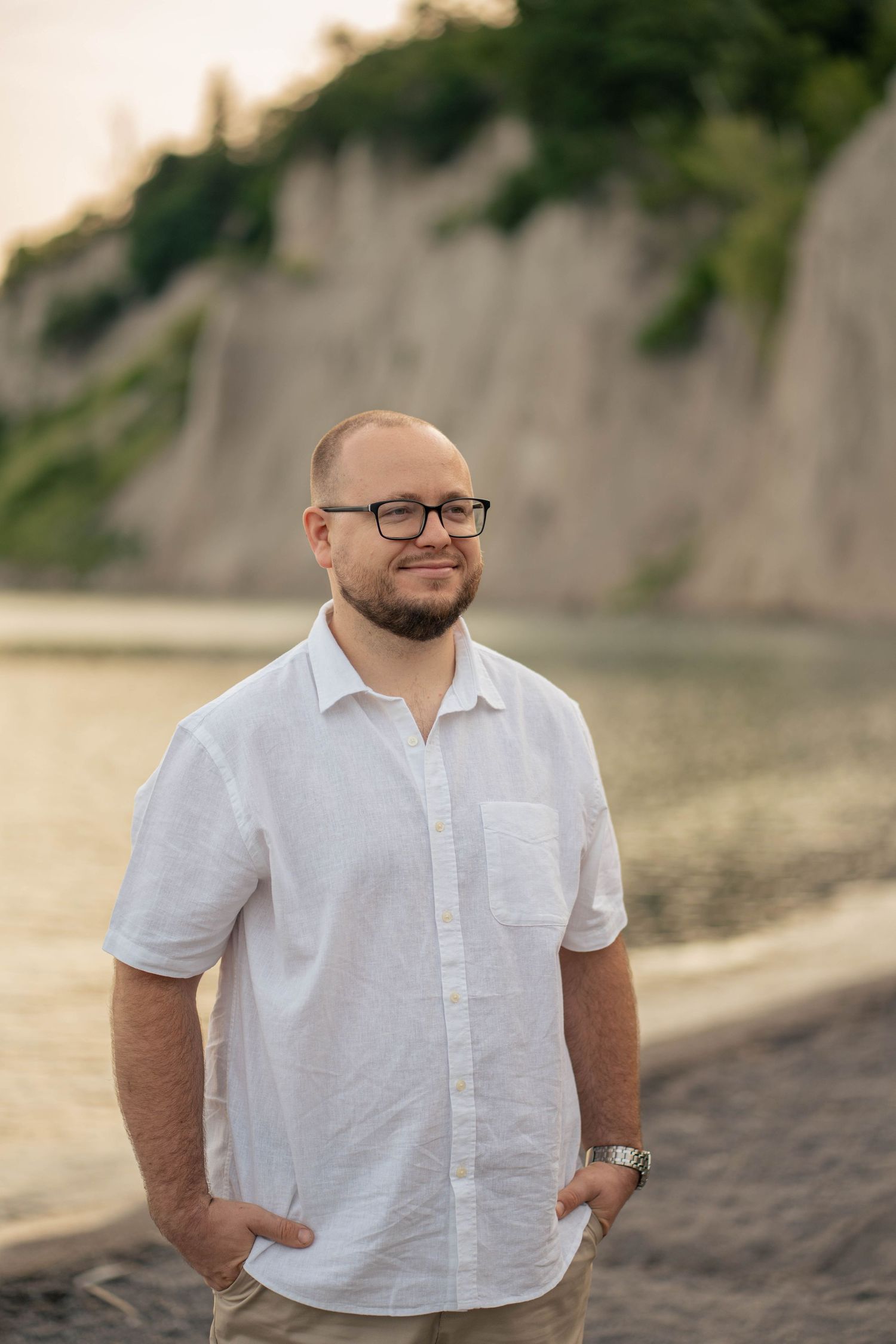 Smiling bearded man with glasses in a white linen shirt poses with hands in pockets near the shoreline and cliffs.