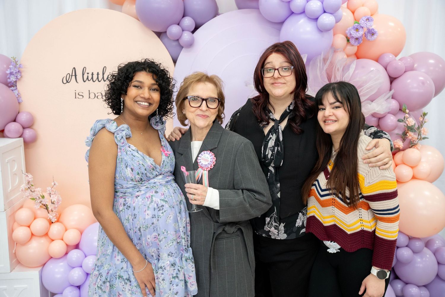 Expecting mother in a floral dress poses with three smiling women at a baby shower decorated with pastel balloons and an arch backdrop.