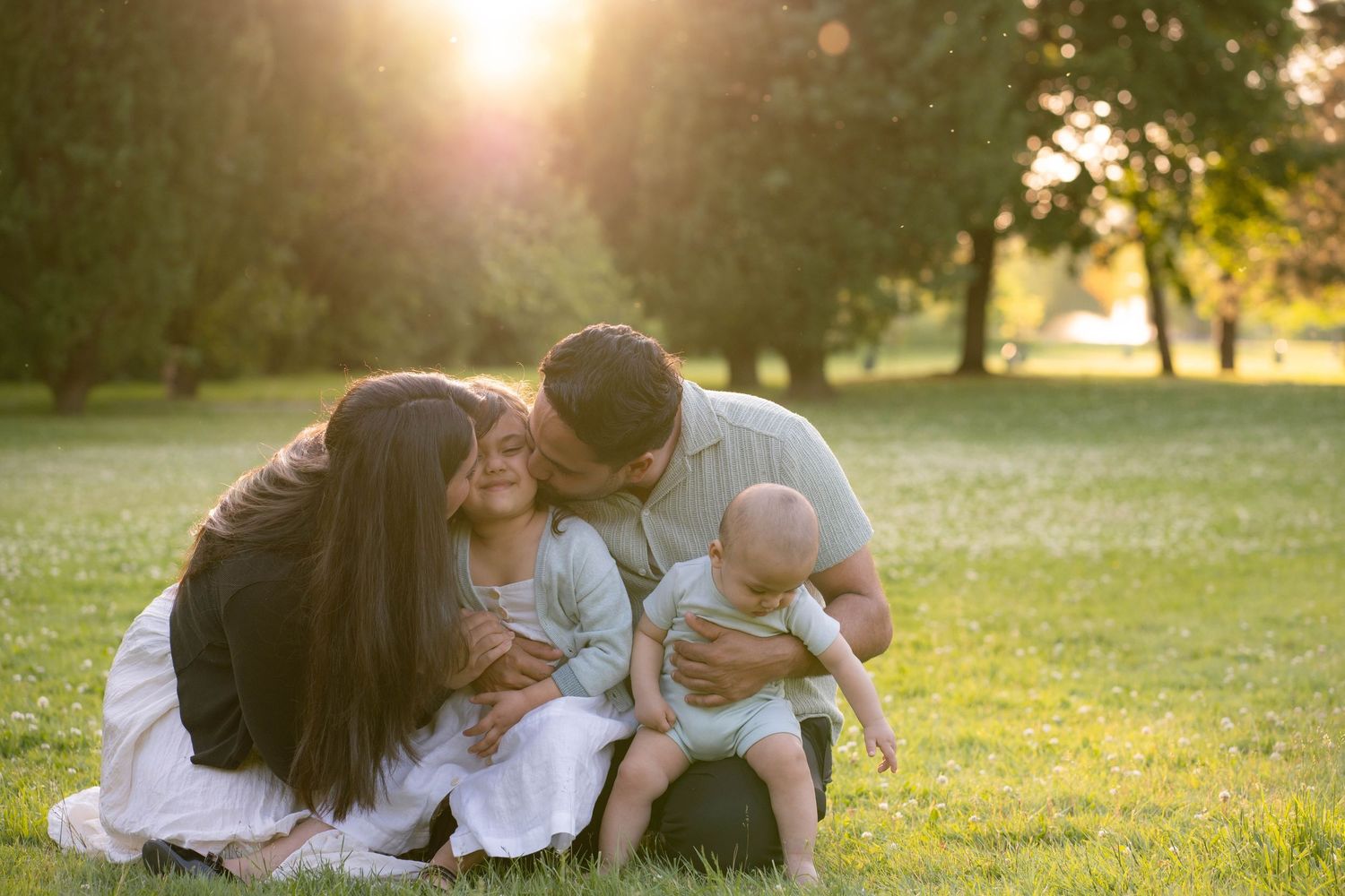 A mother, father, young daughter and baby snuggle together on a grassy lawn in warm golden sunset light.