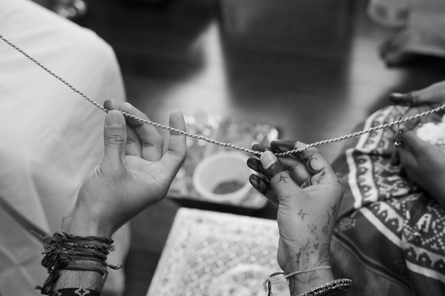 Black and white close-up of two pairs of hands holding a delicate sacred thread during a traditional Indian wedding ritual.