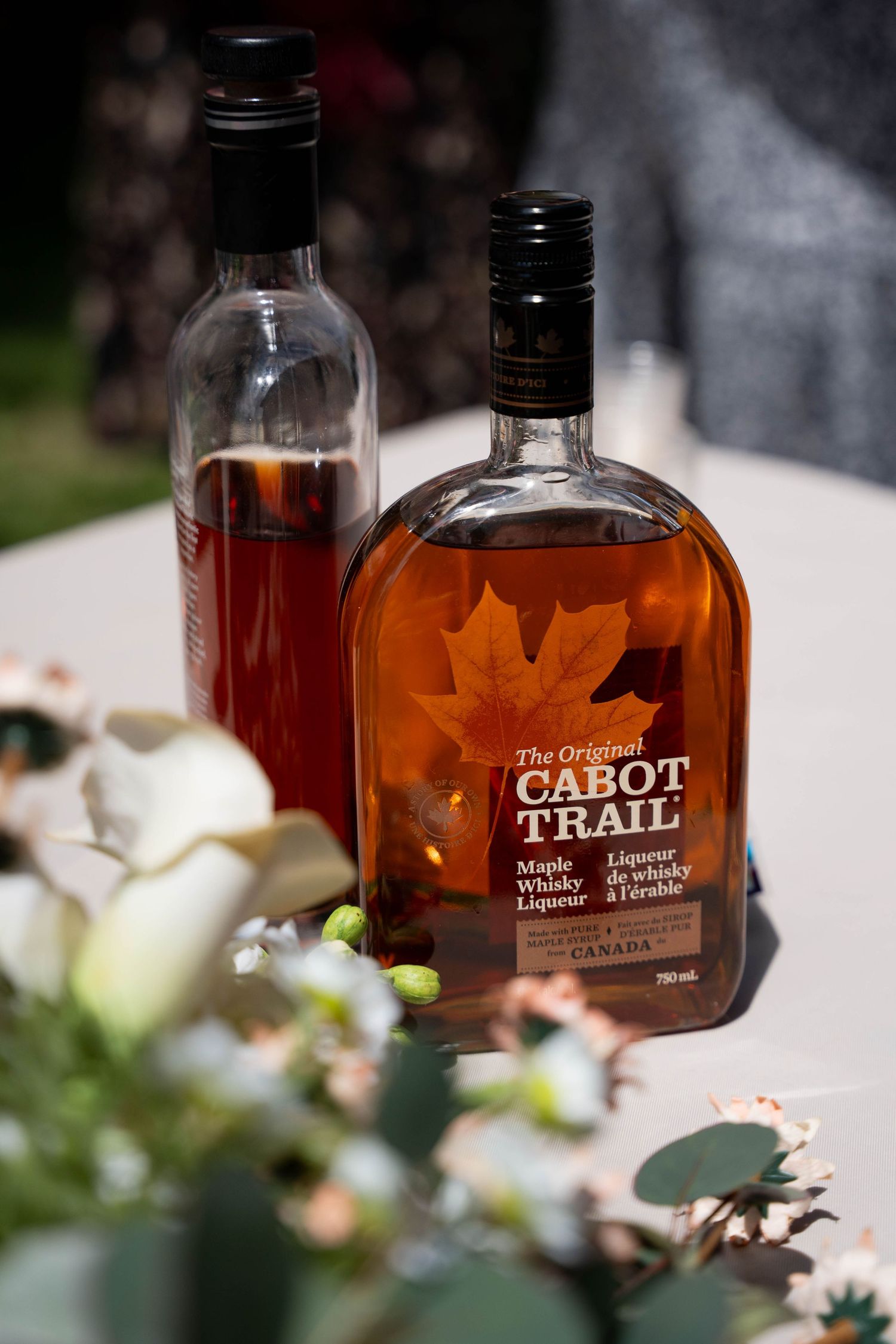 Two bottles of Cabot Trail maple whisky liqueur sit on a table beside white flowers at an outdoor wedding.